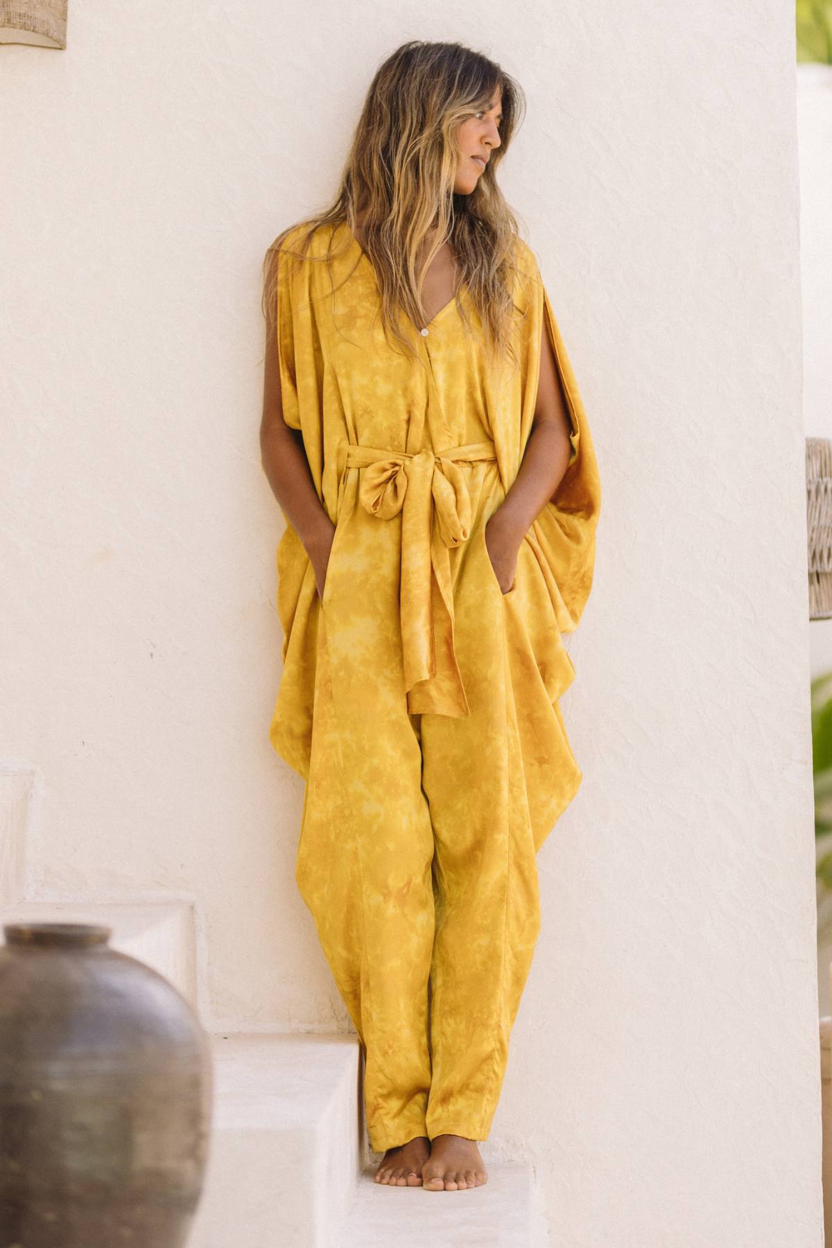 A woman stands on white stairs next to a white wall, wearing the flowing Myrah Penaloza Kuan Yin Playsuit in botanically dyed turmeric gold silk, featuring a tied waist. Her long, wavy hair cascades down as she places her hands in the jumpsuit's pockets while looking to her side. A large, dark jar is partially visible in the foreground.