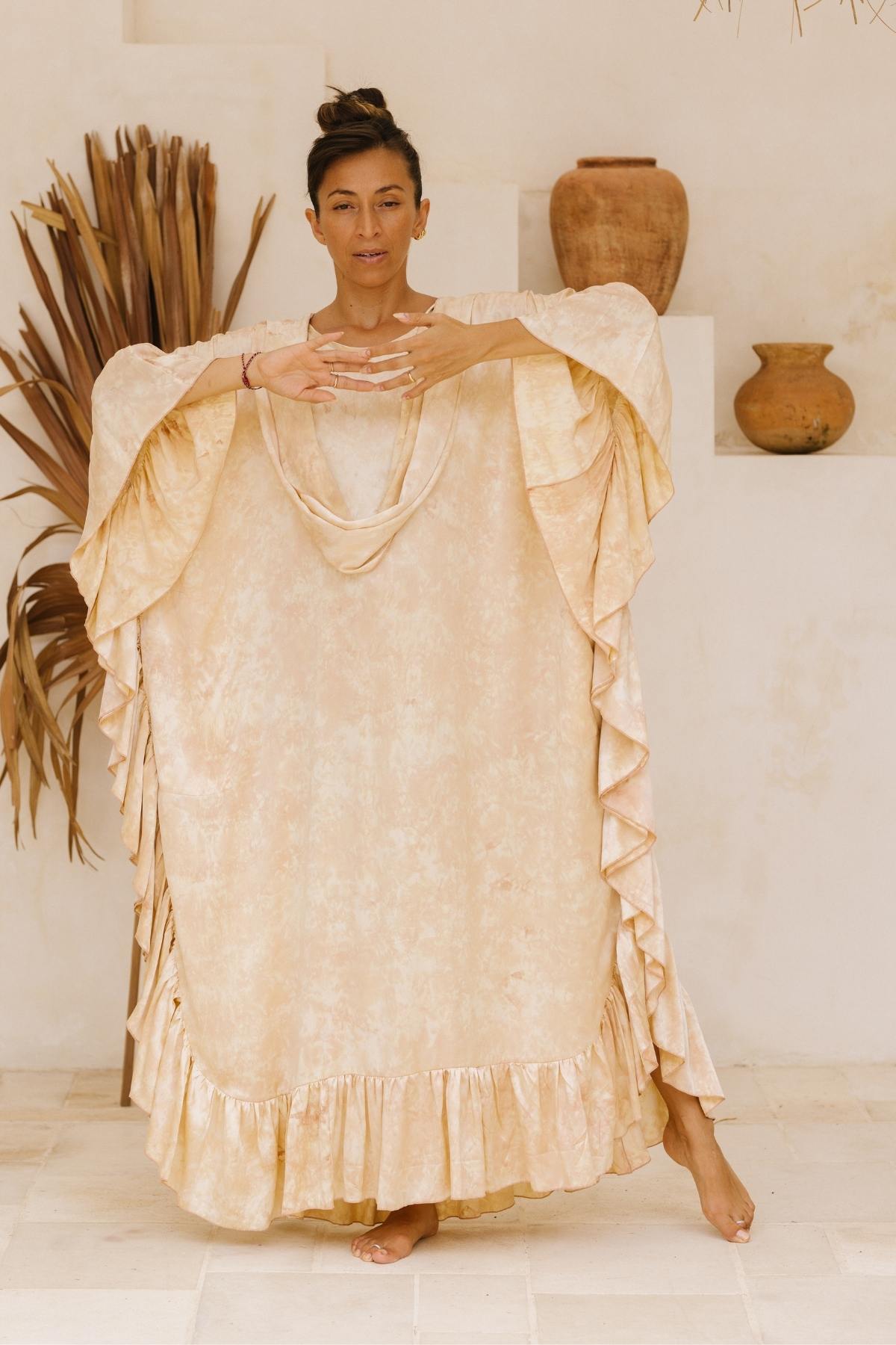 A person dressed in a flowing Perla Silk Gown Made To Order by Myrah Penaloza, characterized by its beige hue and delicate ruffled sleeves and hem, stands on one leg with hands clasped together in a meditative pose. They are positioned against a white wall decorated with vases and dried plants, creating a serene and minimalist ambiance.