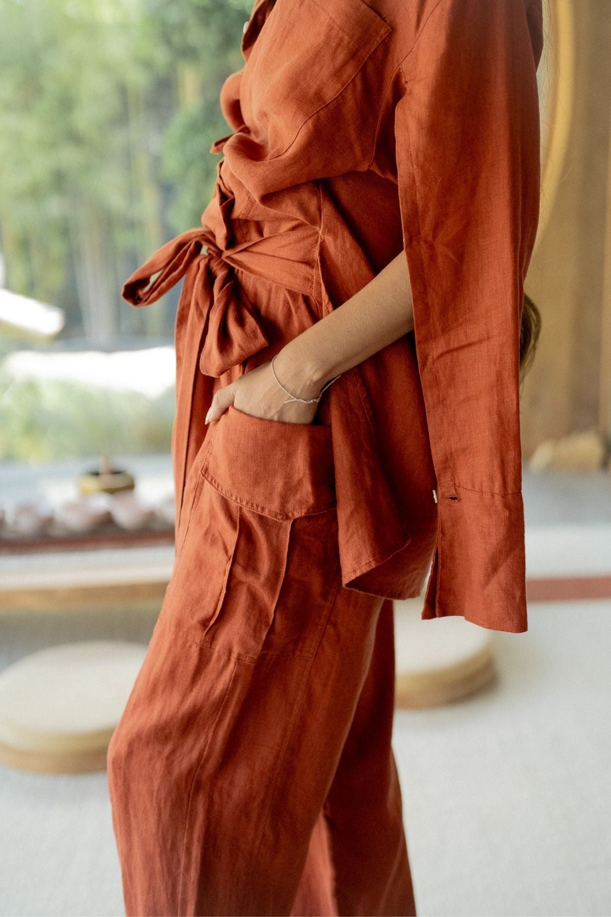 An individual is dressed in the Sutra Linen Set (Made To Order) by Myrah Penaloza, featuring a rust hue, long sleeves, and an integrated belt. They stand with one hand in their pocket against a backdrop of neutral-toned indoor decor and plush furnishings, accentuating the sustainable elegance of their outfit.