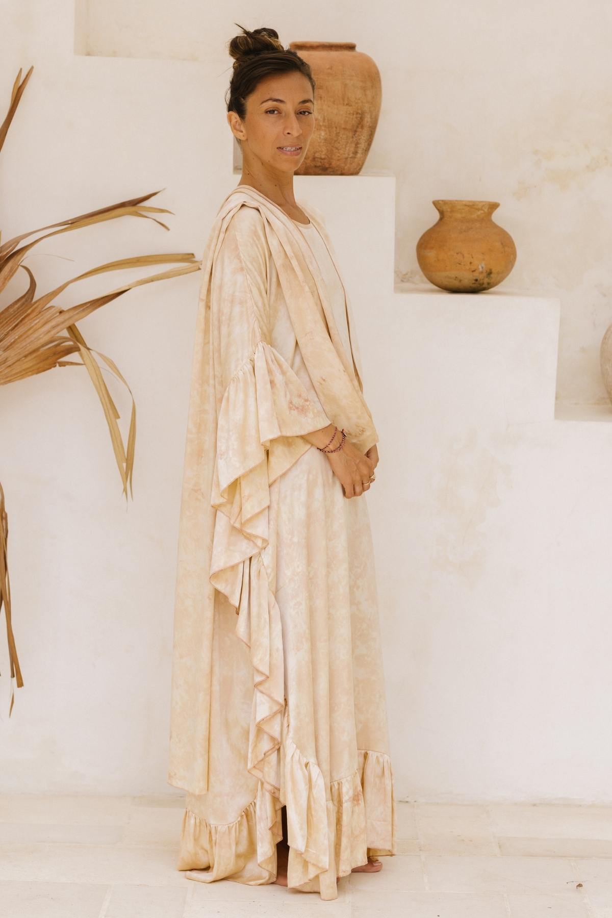 A woman stands sideways in a neutral-toned room, wearing the Perla Silk Gown Made To Order by Myrah Penaloza, a long and flowing beige wedding gown with ruffled edges. Her hair is tied up in a bun, and she has a calm expression. The background features minimalistic decor with clay pots and dried palm leaves, reminiscent of Florence and The Machine's ethereal style.