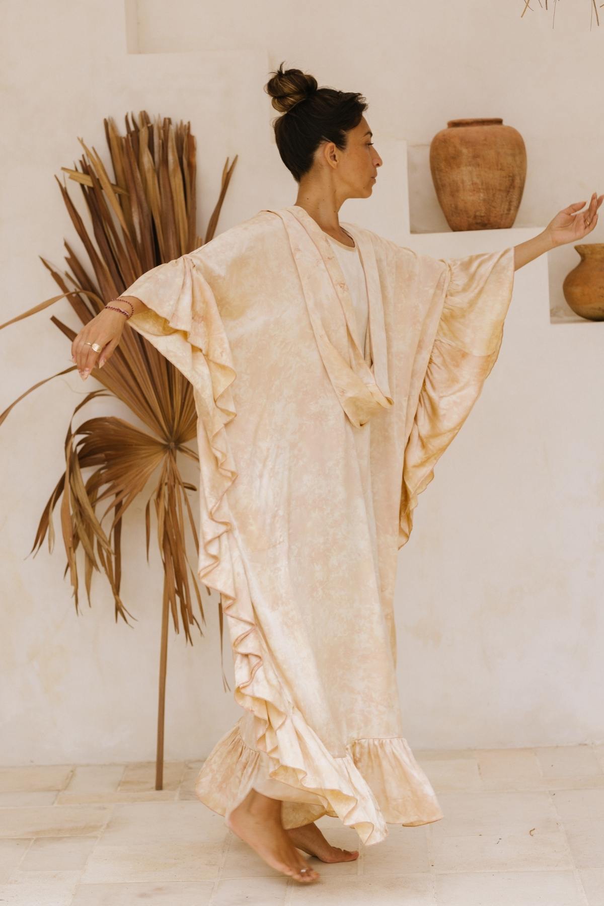 A woman is gracefully dancing barefoot in a light, airy room. She is wearing the Perla Silk Gown Made To Order by Myrah Penaloza, a flowing, cream-colored dress with ruffles and wide sleeves that looks like a custom-made wedding gown. Her hair is tied in a bun, and she is surrounded by decorative dried palm leaves and earthenware pots on shelves as Florence and The Machine plays softly.