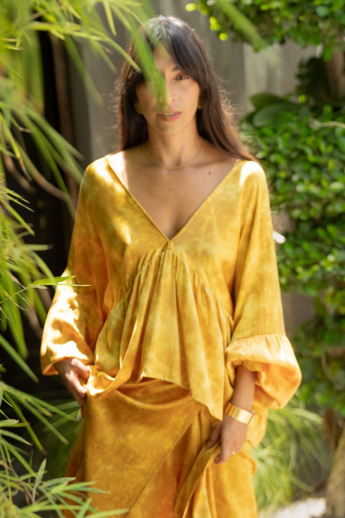 A person with long dark hair stands outdoors, wearing a flowing yellow Shanti Skirt from the Myrah Penaloza Eva Top & Shanti Skirt Set. The background showcases lush green foliage. The scene is softly focused, creating a serene and relaxed atmosphere, enhanced by the elegance of the 100% naturally dyed silk fabric made to order.
