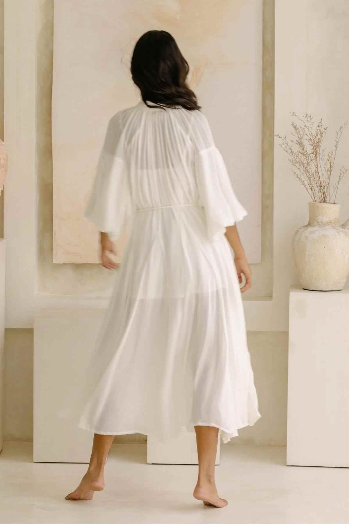 A woman with long dark hair is seen from behind, wearing a flowing Paloma Silk Gown by Myrah Penaloza. She stands barefoot in a minimalist, neutral-toned room adorned with a large vase and dried branches, embodying a bridal-inspired elegance.