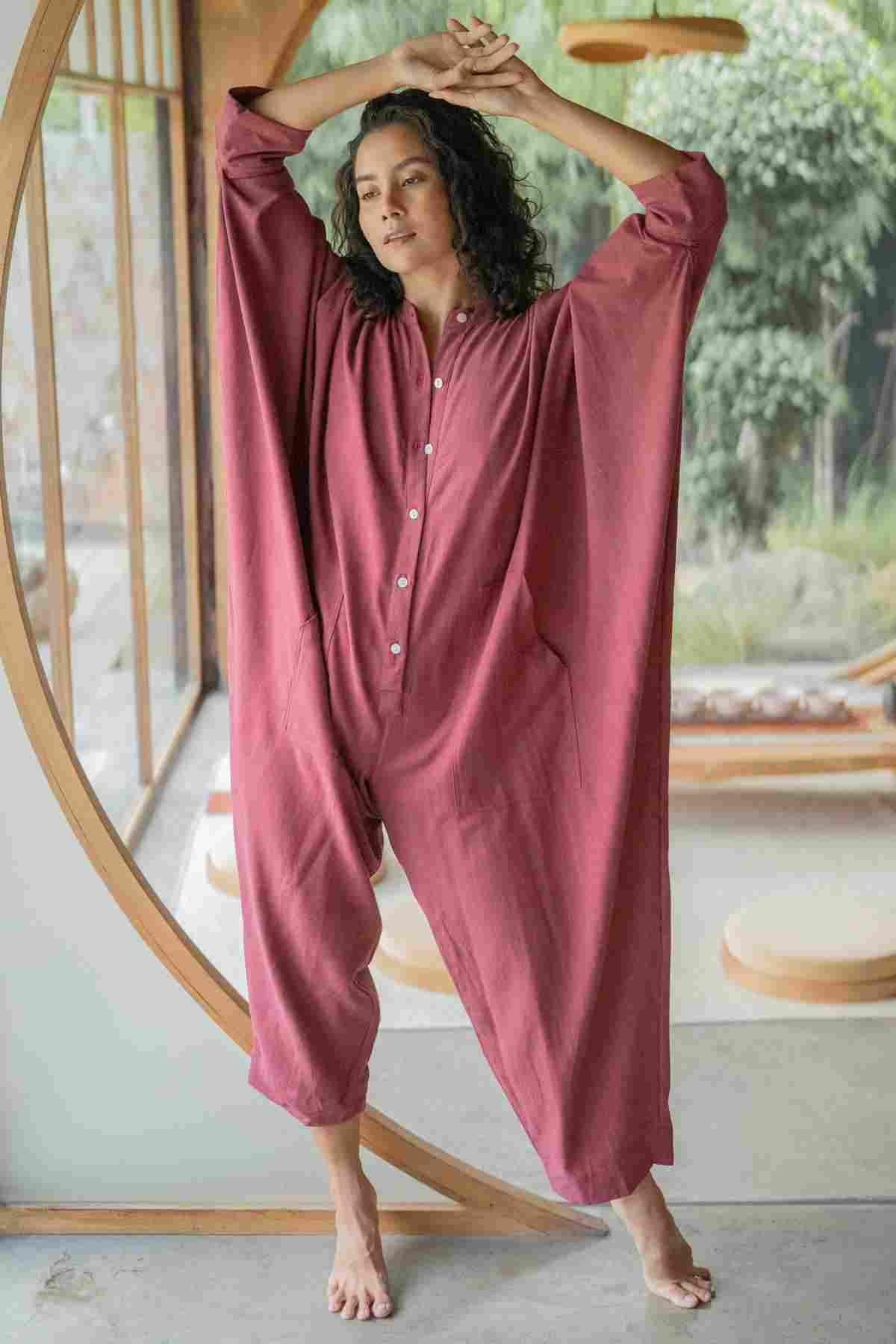 A barefoot person with curly hair stands by a large circular window, stretching with arms above their head. They are wearing a loose-fitting, long-sleeved, rust-colored Miracle Playsuit 2.0 from the limited edition Karama Collection by Myrah Penaloza. The background shows a minimalistic indoor space filled with natural light.