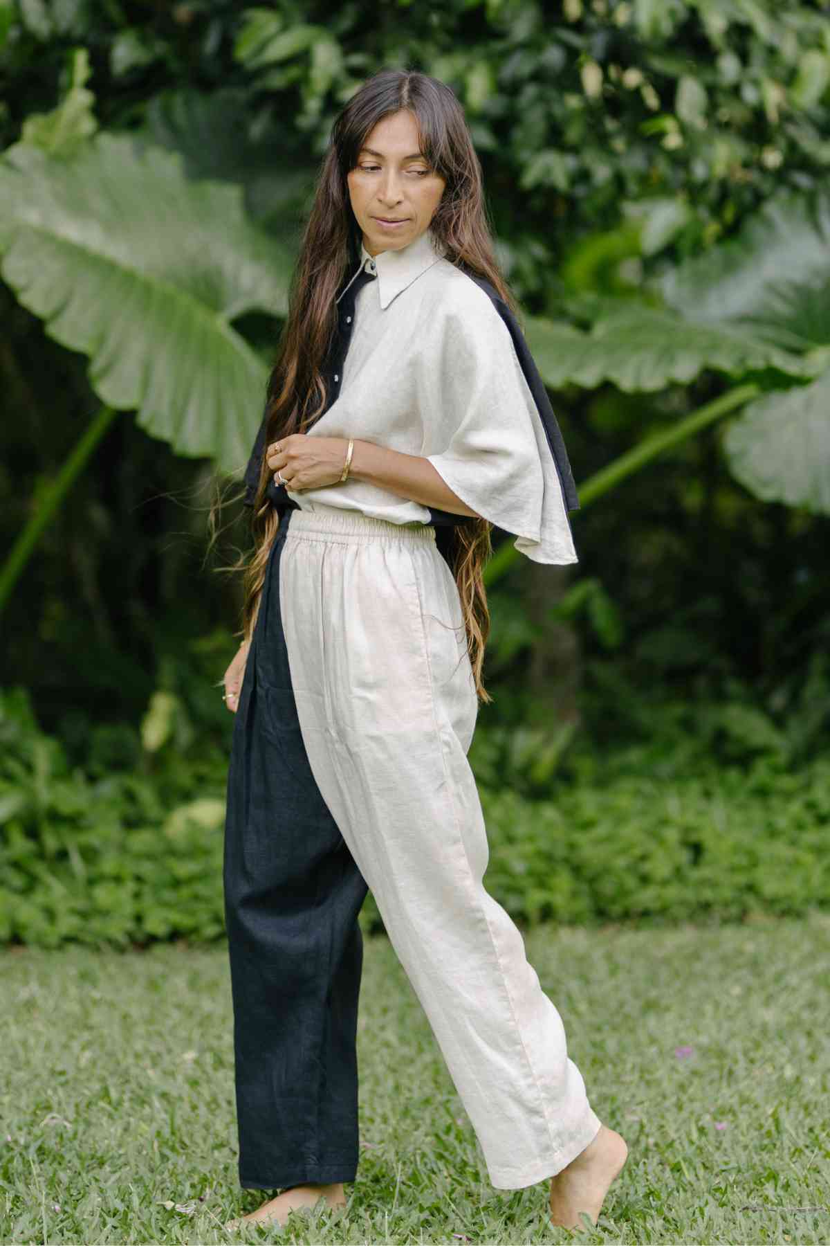A person with long hair is standing barefoot on grass, wearing the black and white Linen Kiki Yin Yang Set from Myrah Penaloza, made of 100% stone-washed linen. Their high-waisted, two-tone pants complement the set perfectly. The background features large green leaves and lush vegetation.