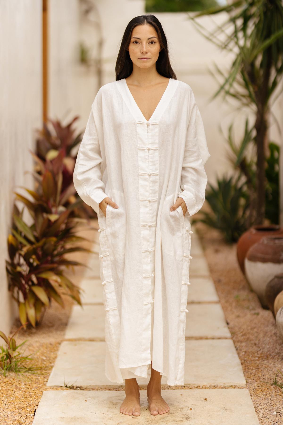 A woman stands barefoot on a gravel path lined with potted plants, wearing a long, loose-fitting white Ancient Chajin Jacket from Myrah Penaloza, made of 100% linen and adorned with Japanese inspired buttons. With her hands in pockets and long, straight brown hair cascading down, she appears calm and serene as she looks directly at the camera.