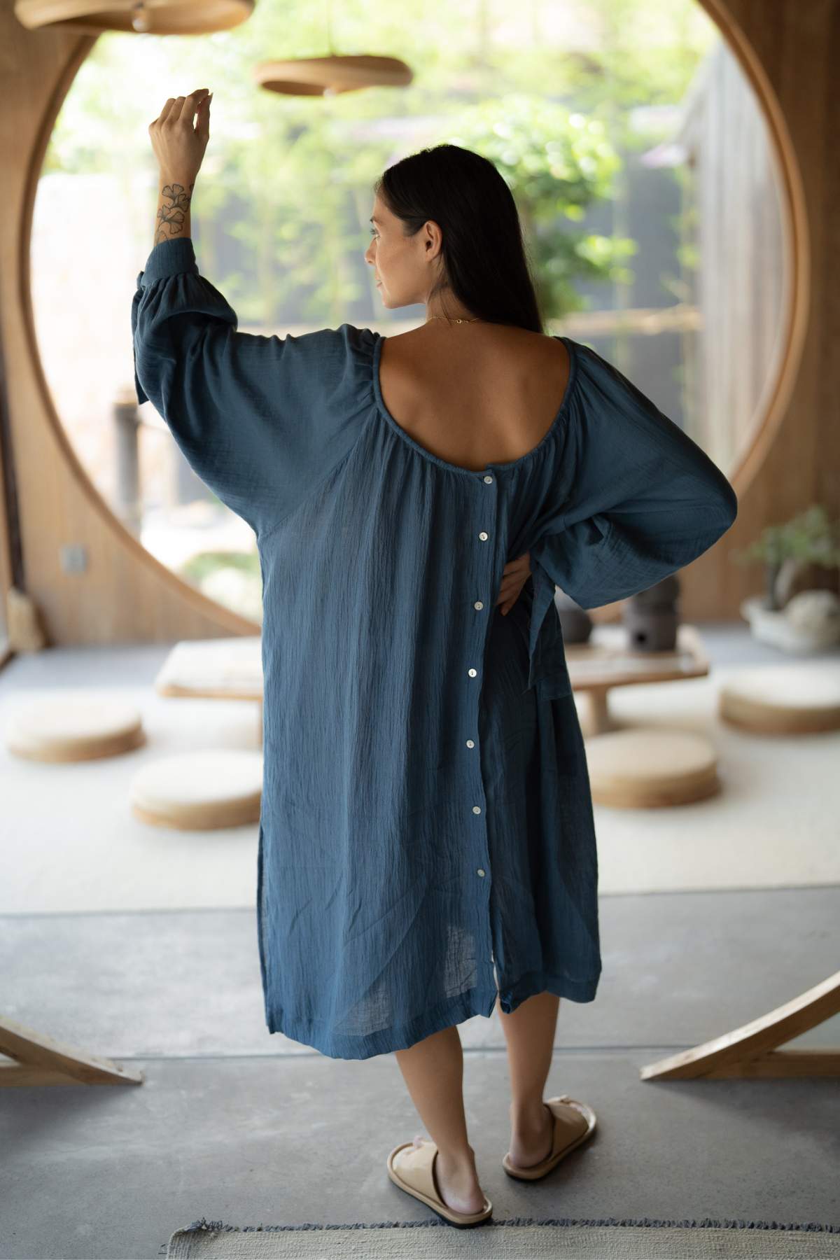 A woman with long dark hair, wearing a Myrah Penaloza Longing Gown in blue linen cotton, stands indoors with her back to the camera, her hand raised. The reversable gown features buttons down the back. The setting includes wooden decor and circular windows, creating a peaceful atmosphere with Virgo details.