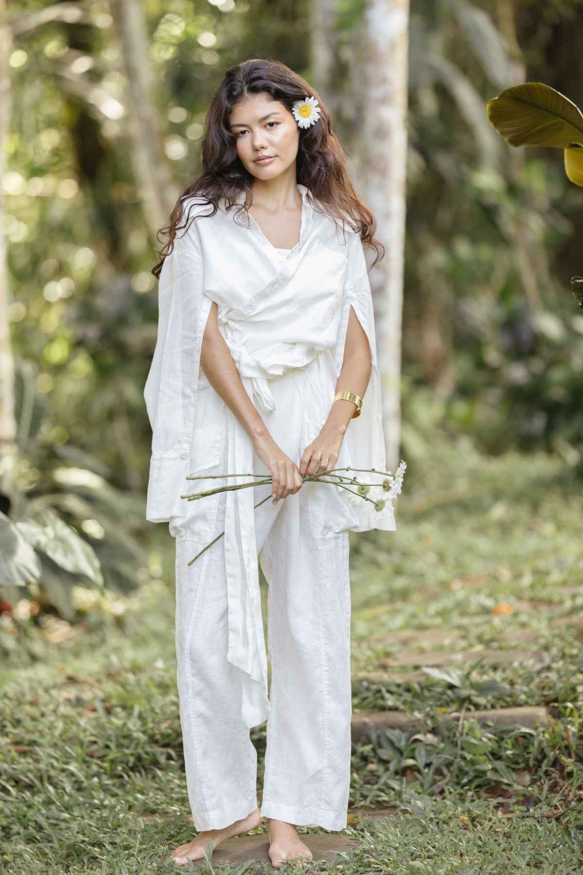 A woman stands barefoot on grass in a serene outdoor setting, donning the versatile Sutra Linen Set by Myrah Penaloza. She holds white flowers, her dark hair loose with a white blossom tucked in it. The flowing outfit complements the lush greenery and trees in the background.