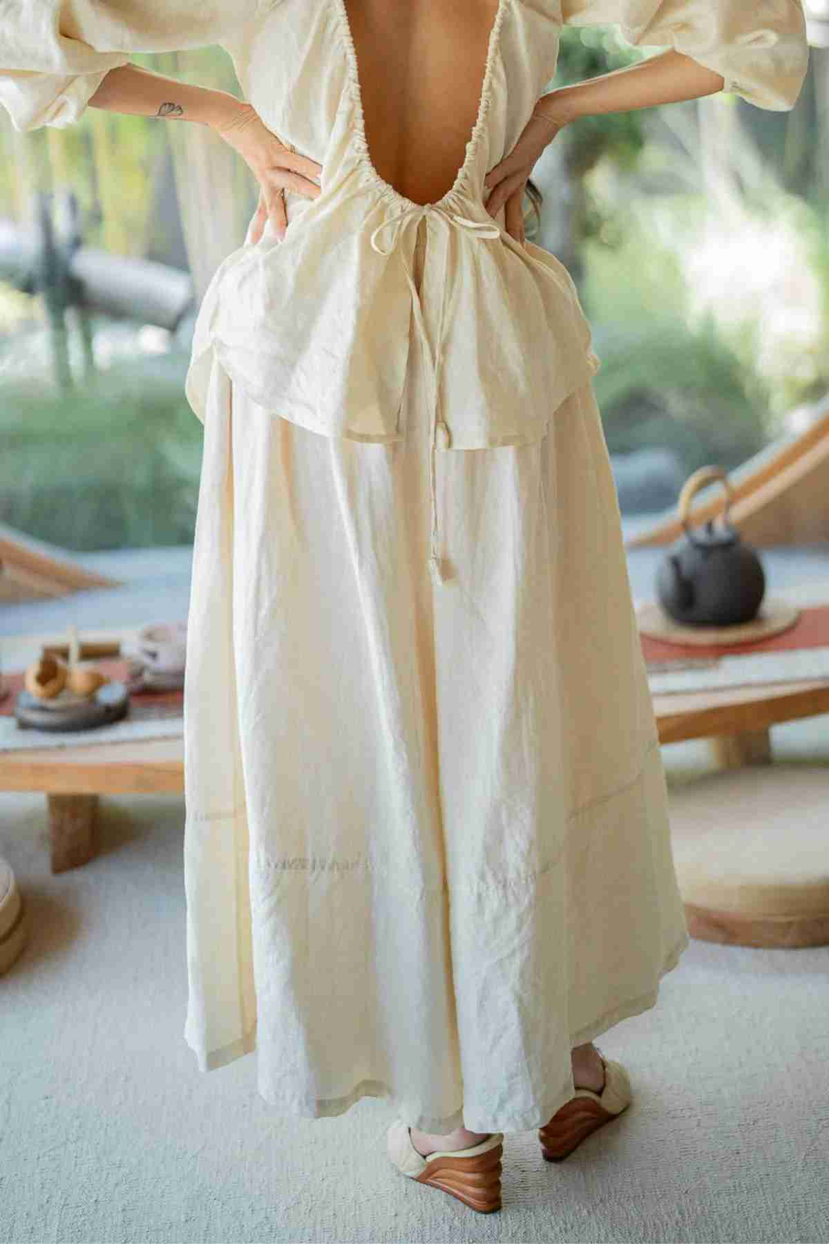 A person confidently stands with hands on hips, wearing a flowing, cream-colored dress with an open back. They are indoors on a light carpet with Pantalon Linen Pants (Wide Legged) by Myrah Penaloza laid over a low wooden table beside them. Greenery visible through the window in the background enhances the graceful setting.