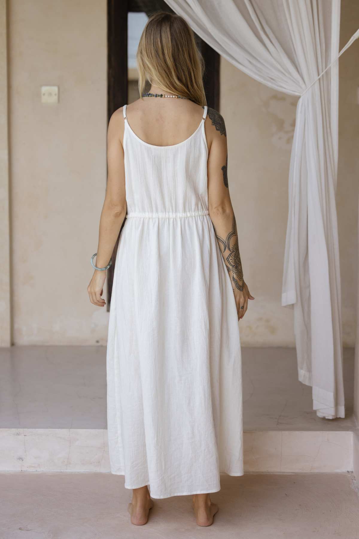 Standing barefoot on a light-colored floor, a woman with long hair emanates effortless elegance in the Myrah Penaloza Dora Slip Dress, featuring a sleeveless design and gathered waist. Her right arm is adorned with tattoos, while a sheer white curtain subtly highlights the timeless piece.