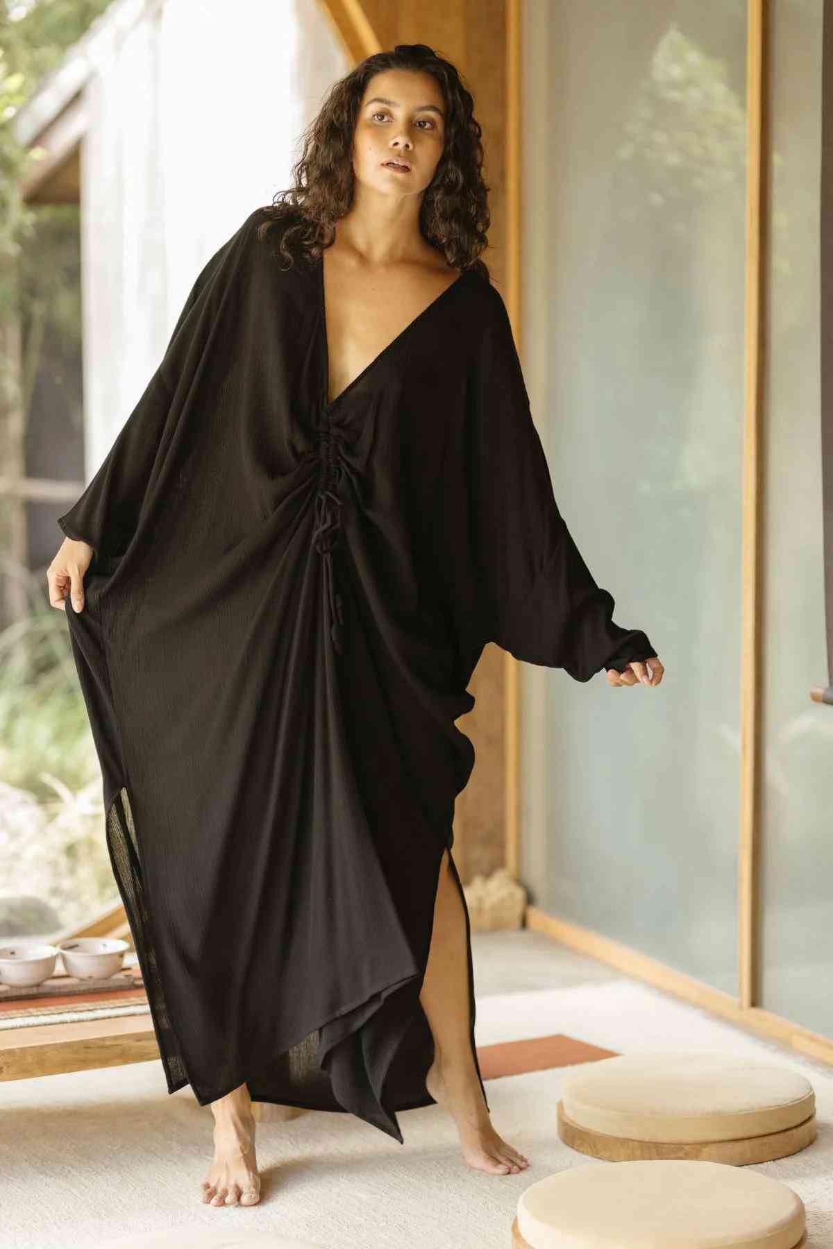 A person with long curly hair, gracefully poised in a flowing Bamboo Rayon Virgo Rising Gown by Myrah Penaloza, stands indoors near large windows with light streaming in. One hand is lifted as if in mid-motion, exuding elegance. The background reveals a view of greenery and minimalistic decor.