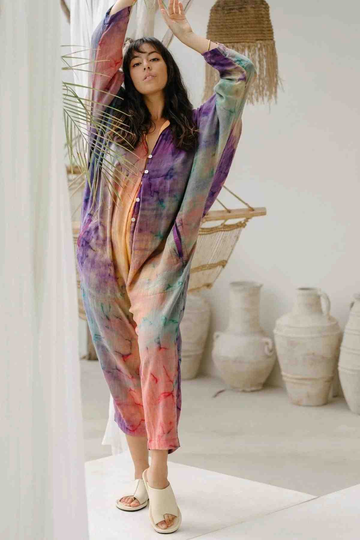A woman with long dark hair stands indoors in a brightly lit room wearing a colorful Sat Torri Rainbeau Playsuit by Myrah Penaloza, which features a retro 1980's design and is handmade. She has one arm raised, touching the back of her head. In the background, there are white clay pots and a hanging woven chair.