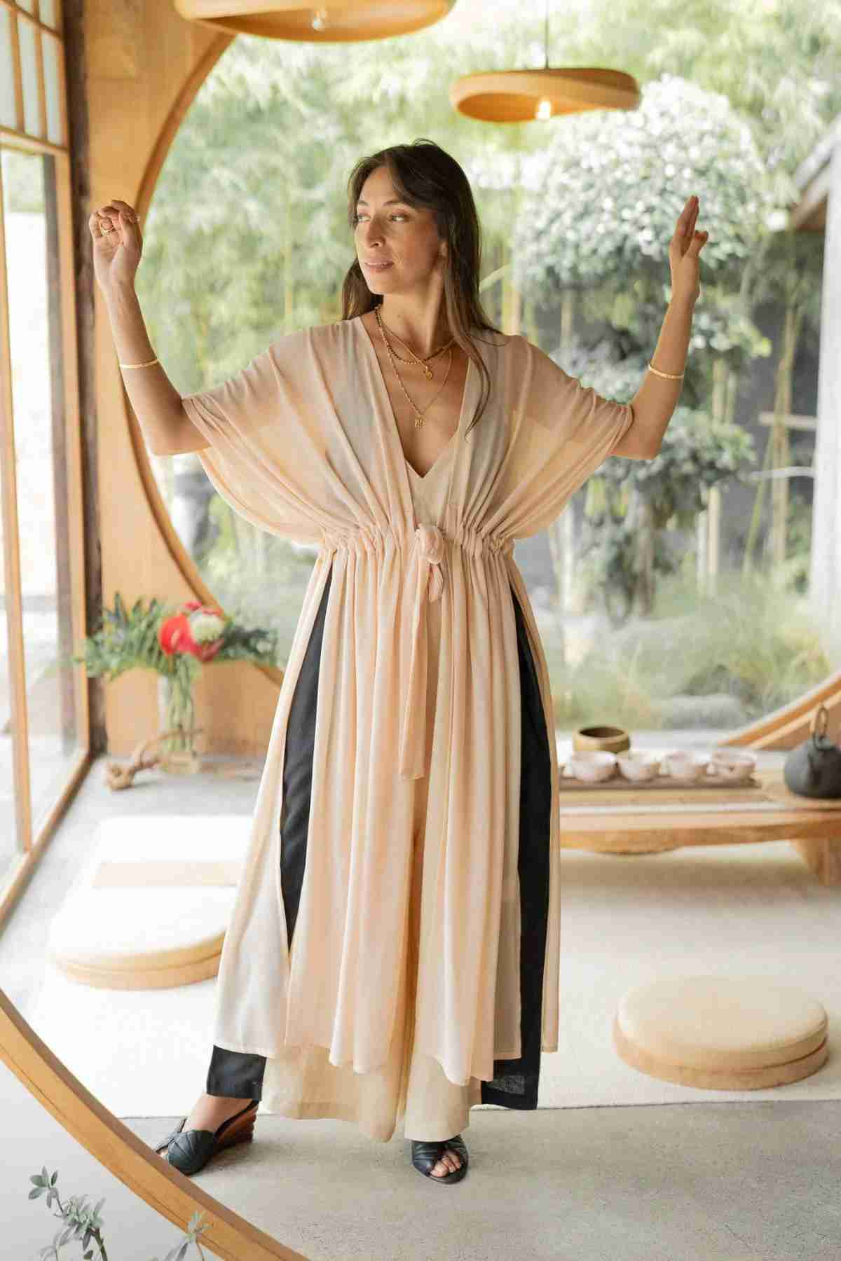 A woman stands in a serene room adorned with circular wooden elements and expansive windows, draped in a flowing Madelin Sheer Chiffon Long Cover by Myrah Penaloza over black pants. With her arms gracefully lifted, a lush green garden is visible in the background, embodying sheer brilliance.