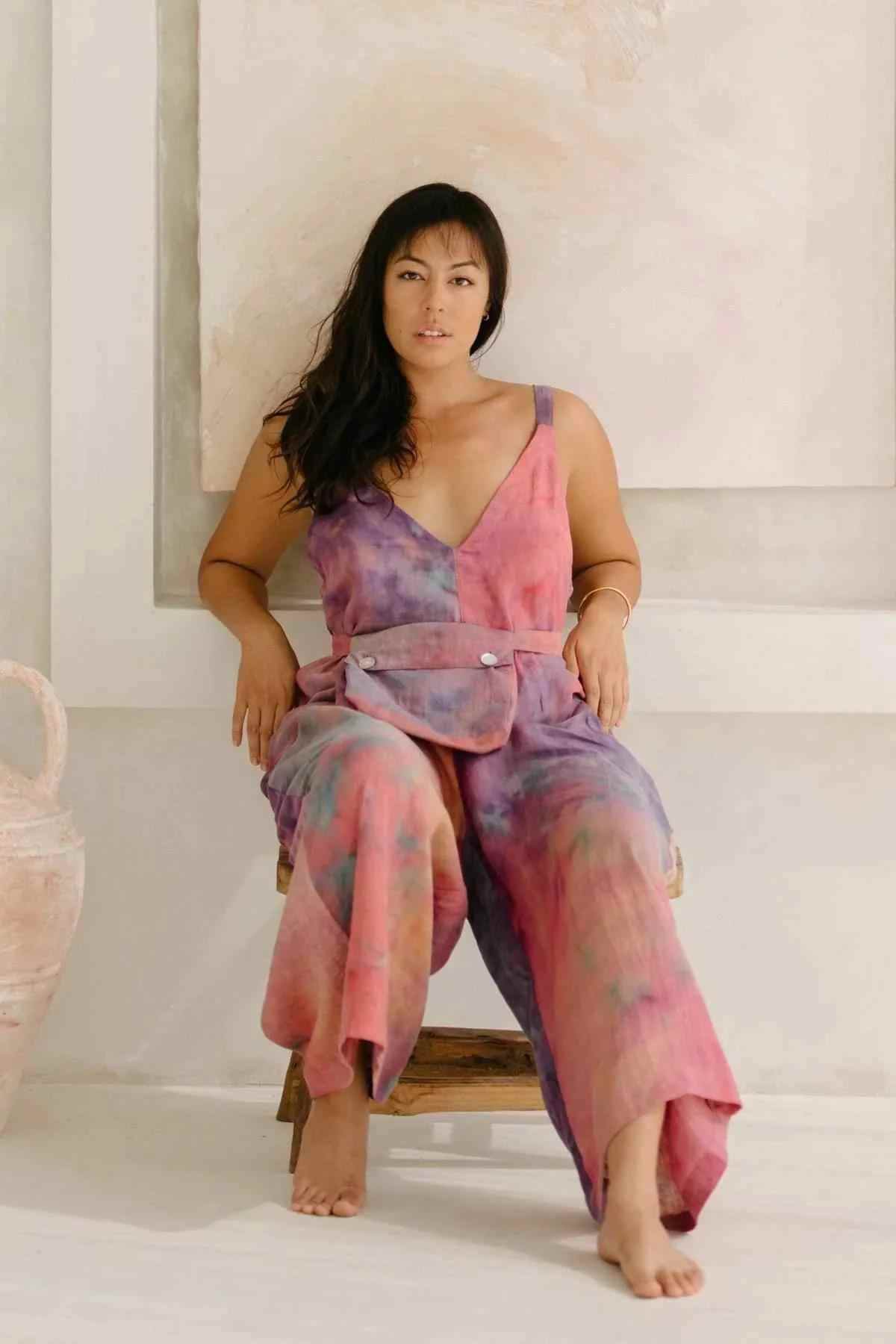 A woman with long dark hair sits on a wooden stool against a neutral-toned backdrop. She is wearing the elegant Rainbeau Linen Kundalini Playsuit by Myrah Penaloza, which features shades of pink, purple, and blue and is cinched with a wide belt. One foot rests on the stool while the other is on the ground.