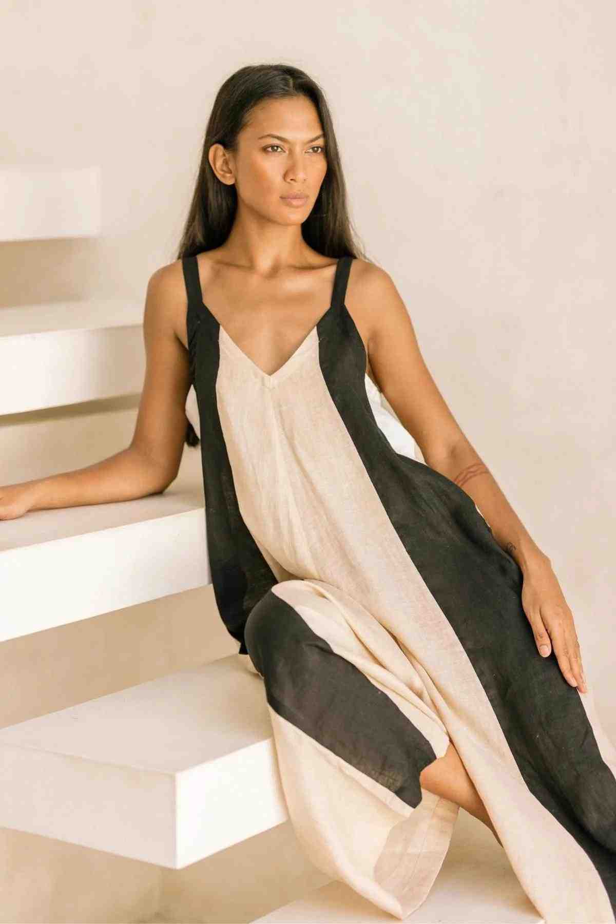 A woman with long, straight hair is seated on white steps. She is wearing the Kundalini Linen Playsuit by Myrah Penaloza, which features a sleeveless design with black and beige vertical panels and wide-leg bottoms. She gazes slightly to the side with a neutral expression.