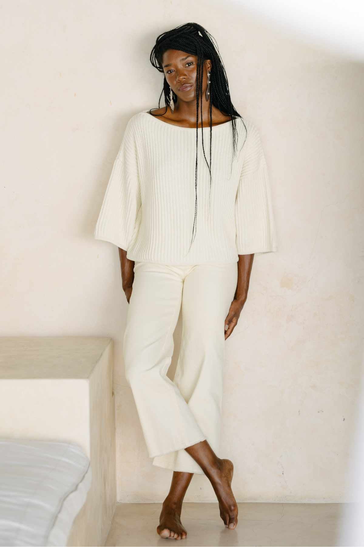 A person with long braided hair stands barefoot against a light-colored wall, gently leaning on their left side. They are wearing the luxurious "Spirit Top Sweater Knit" from Myrah Penaloza, along with matching wide-leg pants. They look serene and relaxed in the minimalist setting.