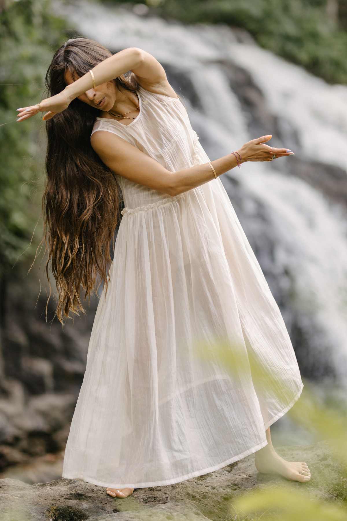 A person with long, flowing hair wearing the Dolly Night Gown (Sheer, 100% Chiffon Silk) by Myrah Penaloza dances gracefully outdoors. They are barefoot, standing on a rock with a waterfall in the background, and pose with one arm covering their face and the other extended outward.