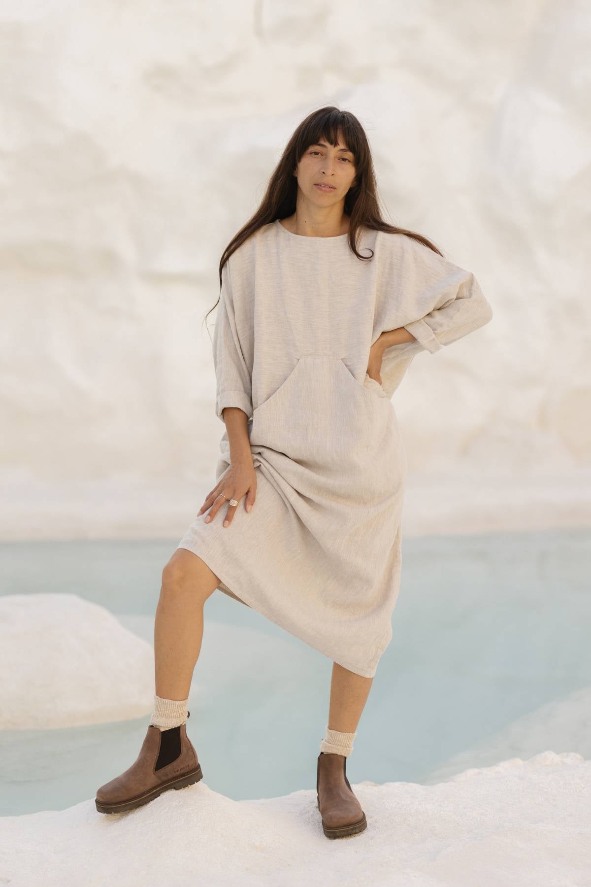 A person stands with one foot on a rock by a pool of water. They have long dark hair and are wearing a loose, light gray Mountain Magic Linen Gown with long sleeves from Myrah Penaloza, paired with brown ankle boots. The background is composed of light-colored rocks or cliffs.