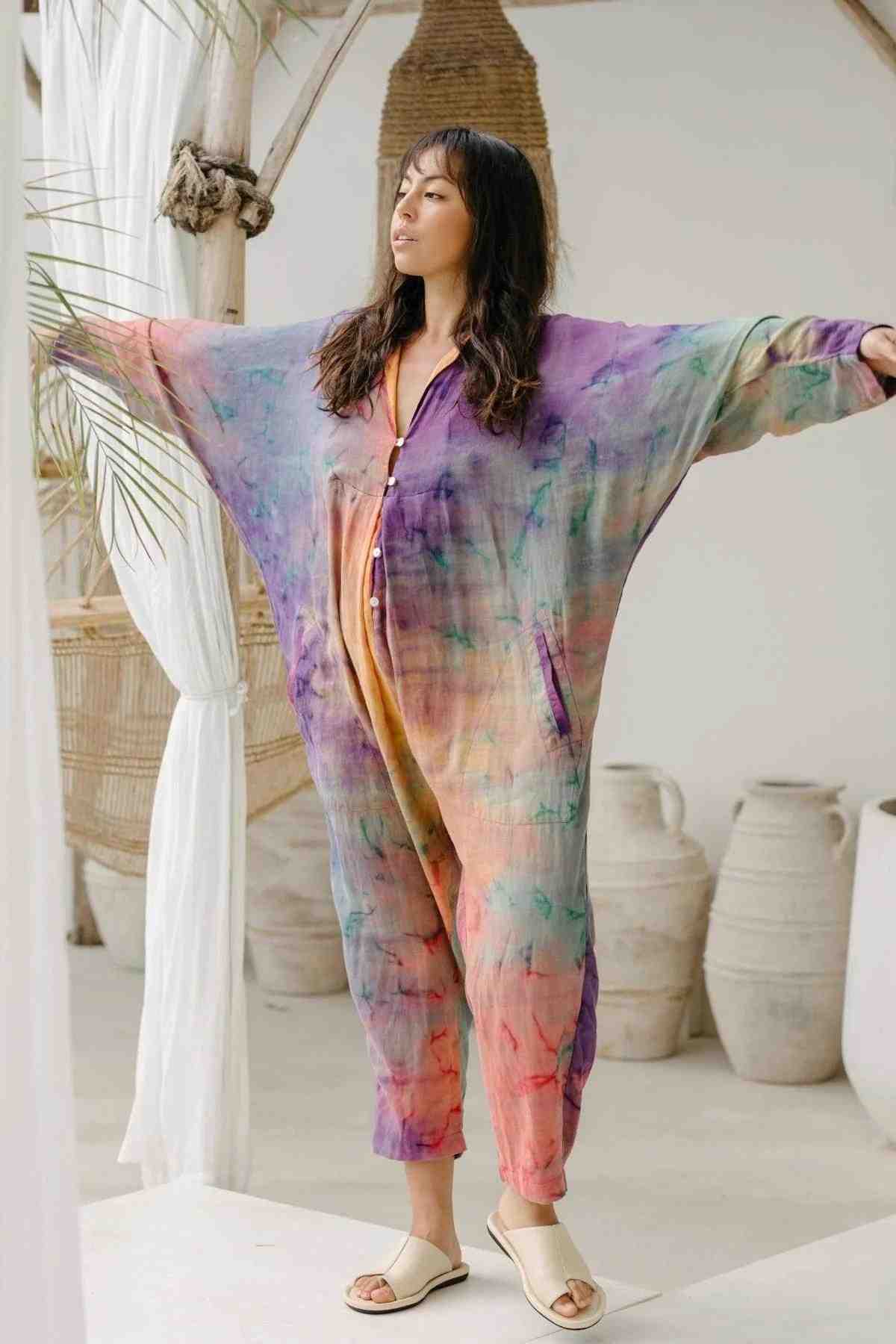 A woman with long dark hair is standing with her arms outstretched, wearing the Sat Torri Rainbeau Playsuit by Myrah Penaloza, a loose and colorful tie-dye jumpsuit inspired by a retro 1980's design. The background features light-colored decorations and large ceramic pots. She is also wearing light-colored slip-on sandals.