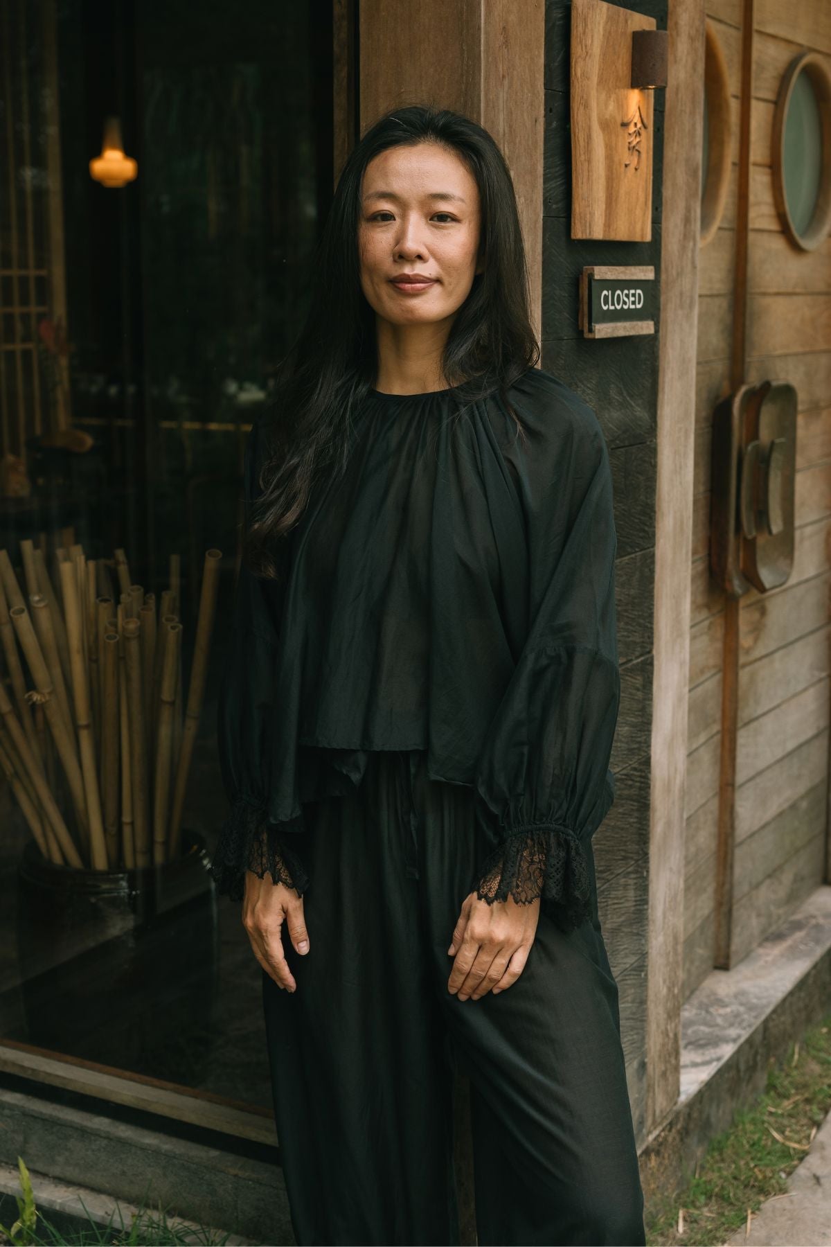 Woman standing at a wooden doorway in Bali wearing the Anusara Evening Set by Myrah Penaloza, a silk and cotton top and pant in Dark Moon Black with hand woven lace at the cuffs.
