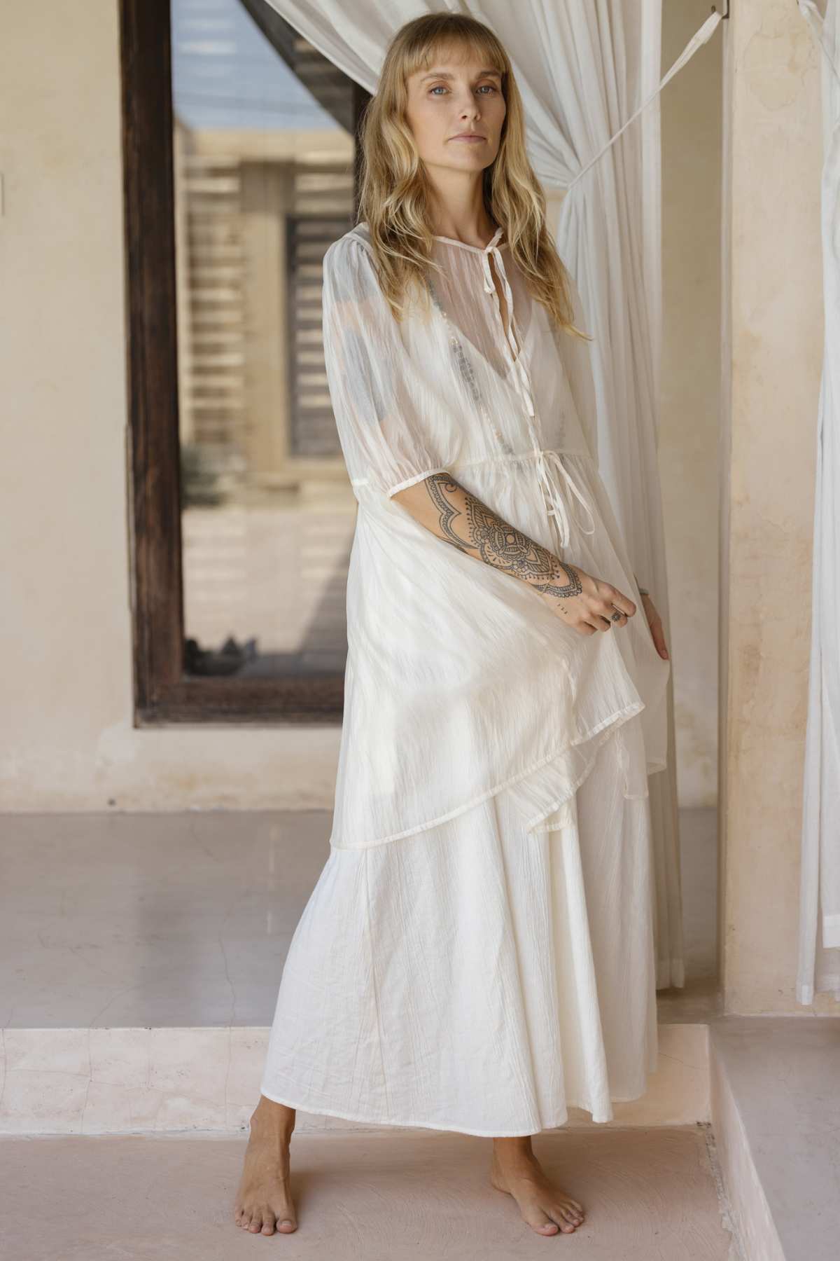 In a neutral-toned room, a woman radiates effortless elegance as she stands barefoot in a flowing Dora Slip Dress by Myrah Penaloza, featuring sheer sleeves. Her long blonde hair gracefully falls over her tattooed forearm while she stands near a window dressed with sheer white curtains. The minimalist setting accentuates an aura of timeless peace.