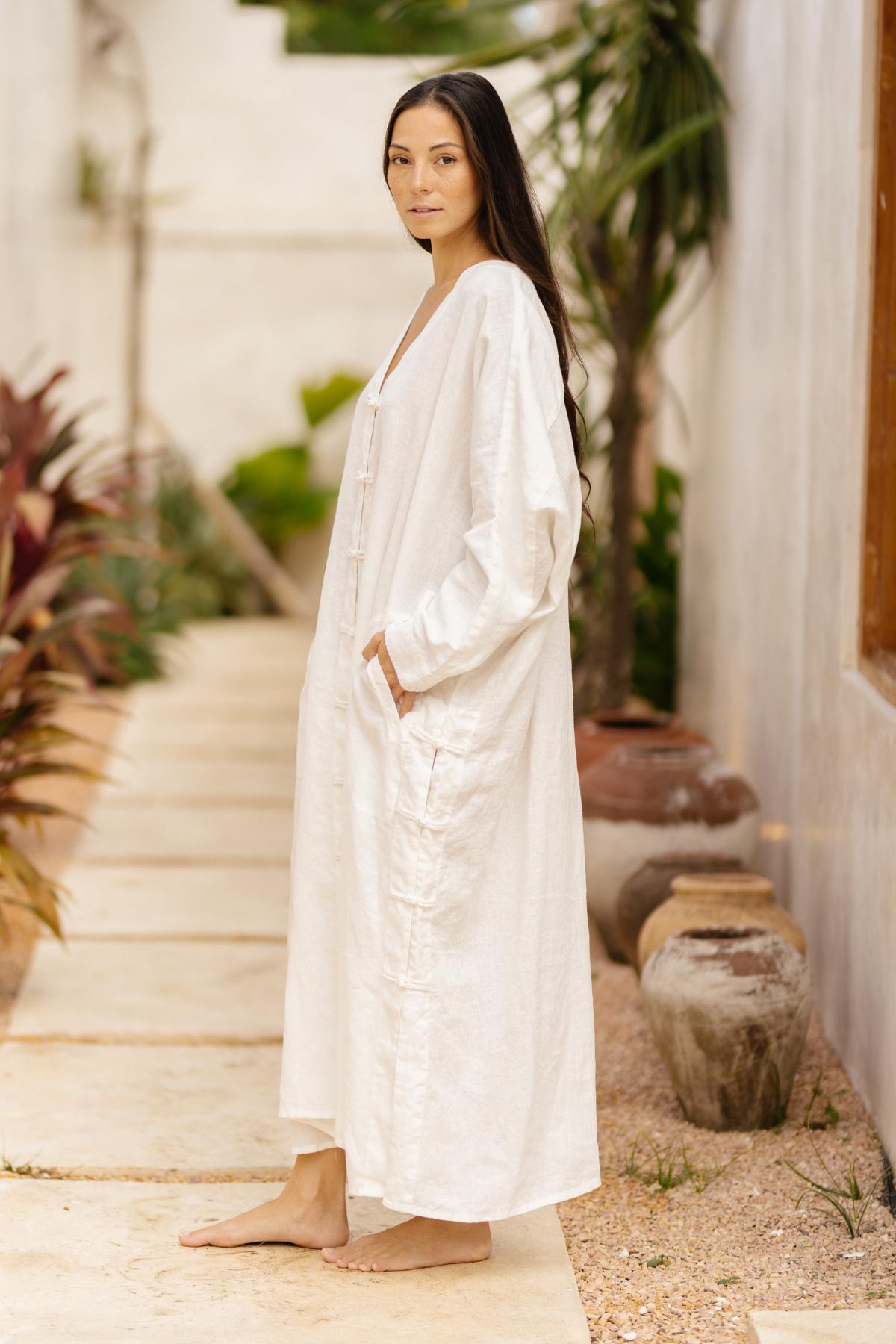 A woman with long dark hair stands barefoot on a stone pathway, wearing the Ancient Chajin Jacket (100% Linen)- Made To Order from Myrah Penaloza. She is surrounded by potted plants and greenery, with a white wall and large window to her right. The setting appears calm and natural.