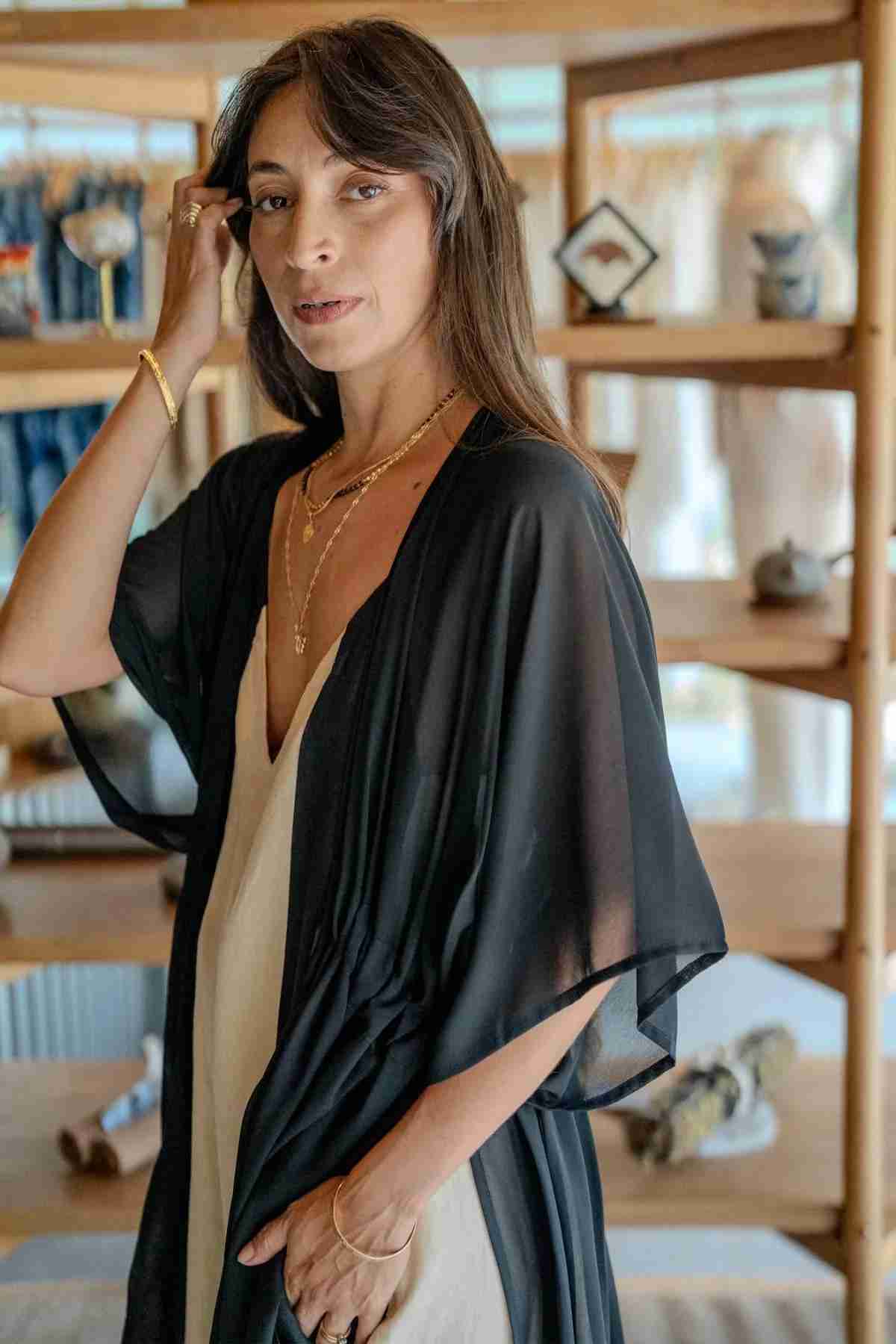 A woman with long brown hair stands inside a stylish, wood-accented store, wearing the Madelin Sheer Chiffon Long Cover by Myrah Penaloza over a light-colored dress, gold necklaces, and bracelets. She is adjusting her hair with one hand while looking towards the camera with sheer brilliance in her calm expression.