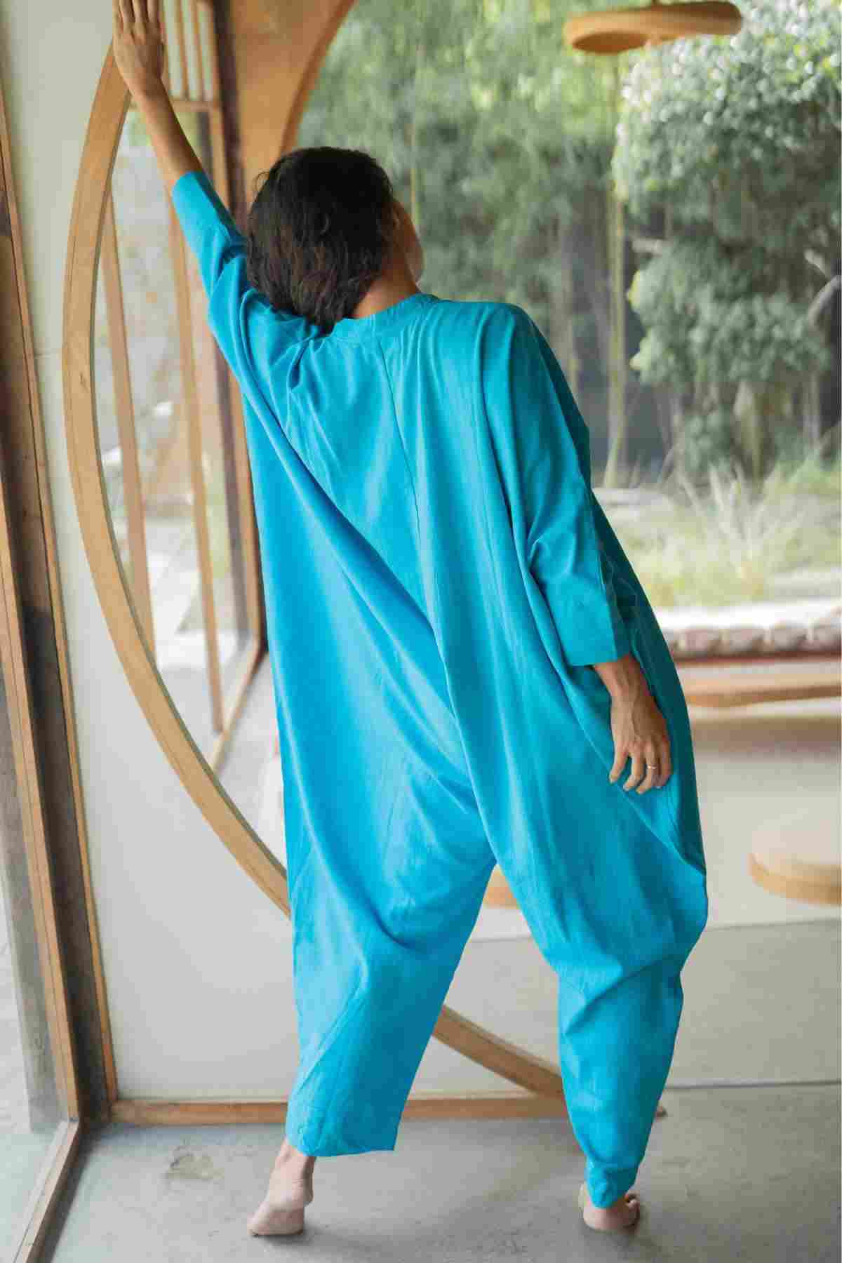 A person in a loose, bright blue Miracle Playsuit 2.0 from the Karama Collection by Myrah Penaloza stands with their back to the camera, one arm extended upward and hand resting on a circular wooden structure. They are in a room with large windows that look out to a lush green garden. This piece is part of our limited edition collaboration collection.