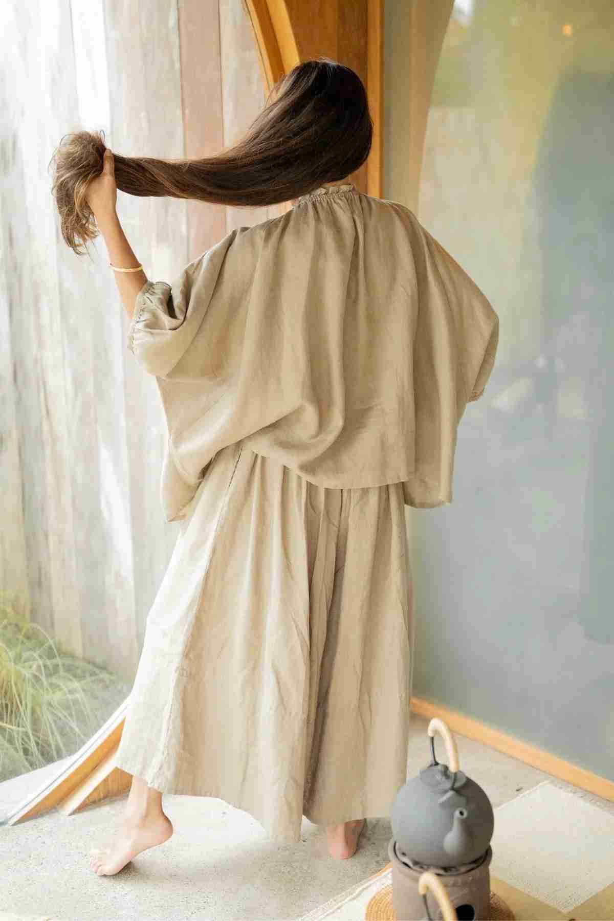 A woman stands barefoot on a wooden floor, facing a window. She wears wide-legged pants from Myrah Penaloza called Pantalon Linen Pants. With one hand, she gently pulls back her long brown hair. The room features curved wooden elements and is illuminated by soft natural light.