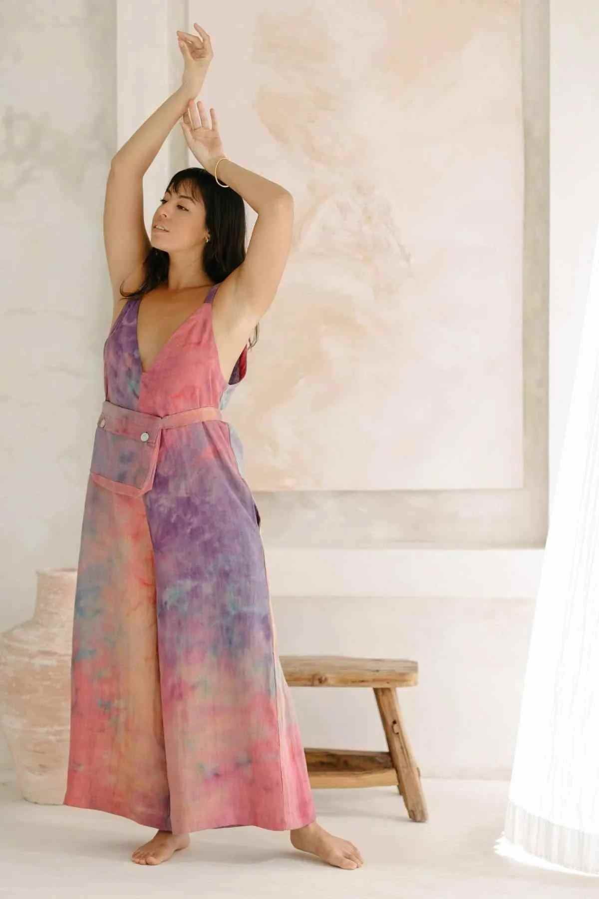 A woman stands barefoot with her arms gracefully lifted overhead, wearing the elegant Rainbeau Linen Kundalini Playsuit by Myrah Penaloza in shades of pink, purple, and blue. She stands inside a minimalist room with a wooden bench and a light-colored wall, as natural light filters through a curtain.
