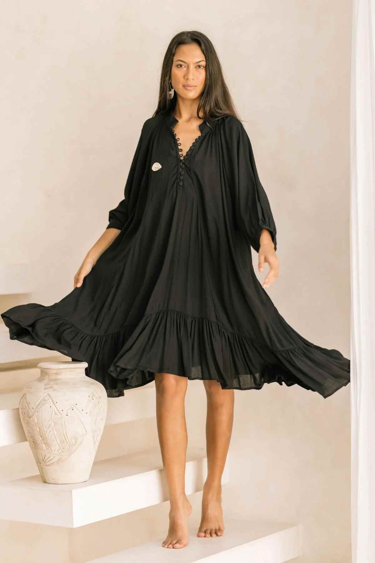 Wearing the Bamboo Rayon FRIDA Gown Short by Myrah Penaloza, a person with long, dark hair stands barefoot on indoor steps alongside an ornate white vase. The flowy black dress is complemented by ruffles at the hem, enhancing the minimalist and elegant decor of the setting.