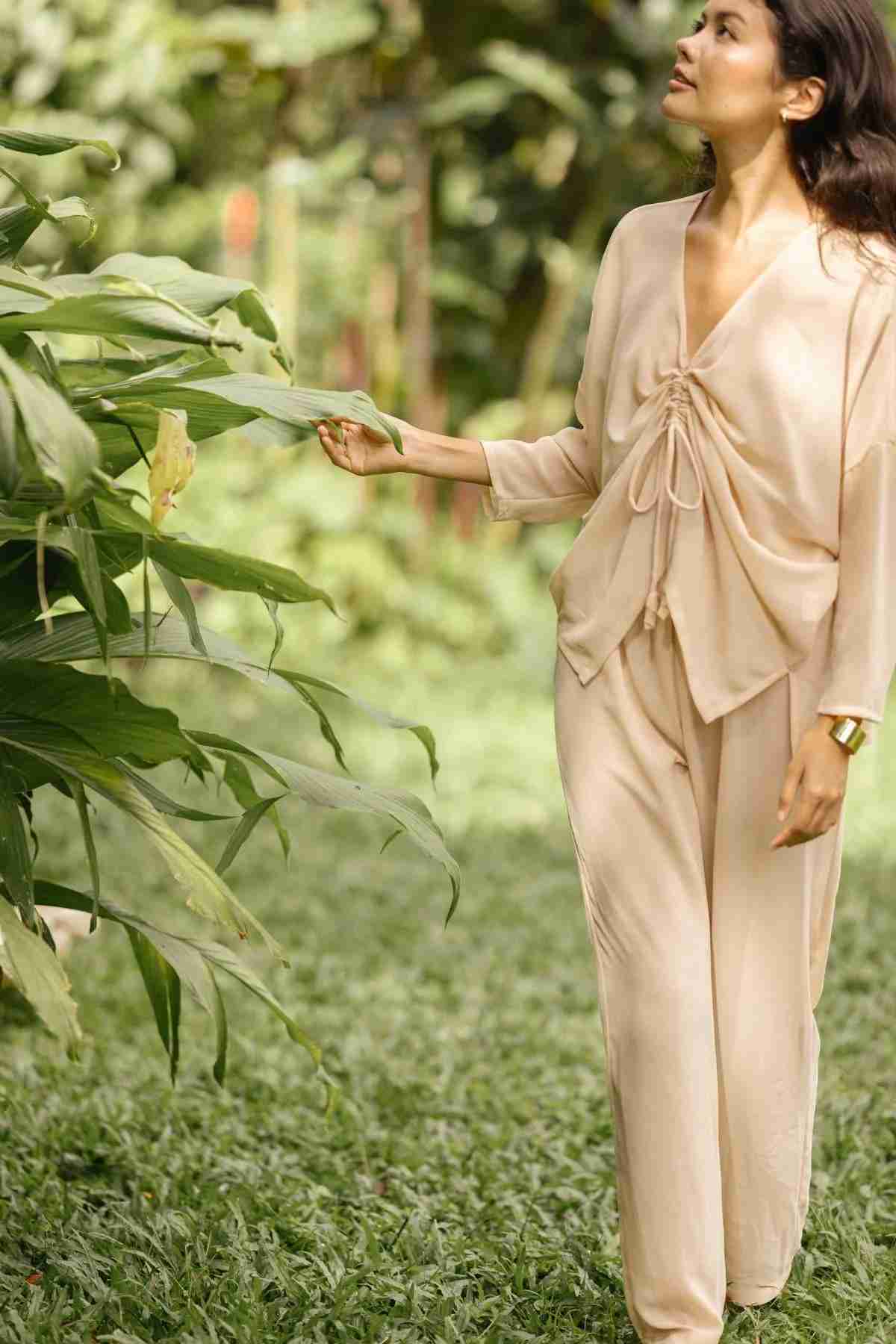 A person in a Chiffon Silk Sheer Virgo Rising Set (Made to Order) by Myrah Penaloza walks through a lush garden, gently touching large green leaves while looking upward. The background features vibrant greenery and sunlight filtering through the trees, creating a luxurious atmosphere.