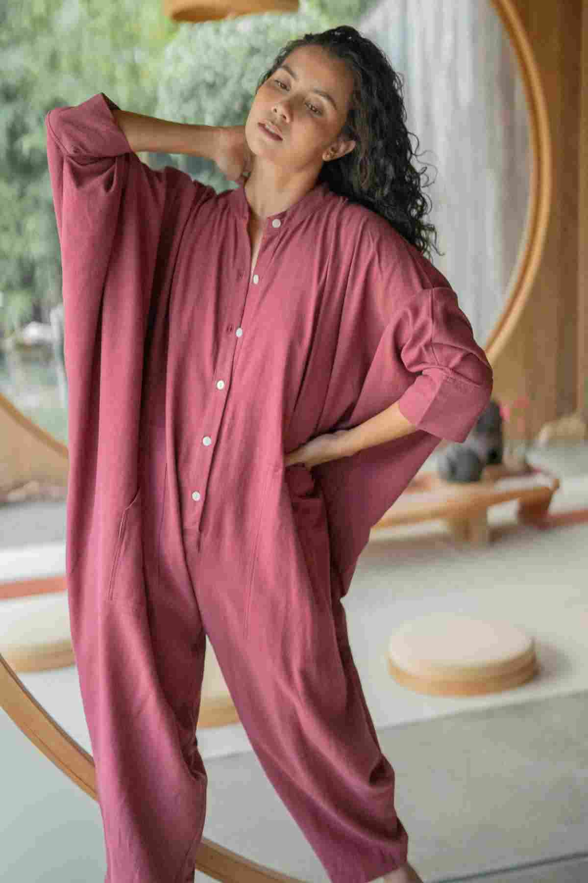 A person stands indoors wearing the limited edition loose red Miracle Playsuit 2.0 from the Karama Collection by Myrah Penaloza, characterized by its large sleeves and front buttons. One of their hands is in a pocket while the other rests behind their head. In the background, circular wooden architecture and a green outdoor scene are visible.