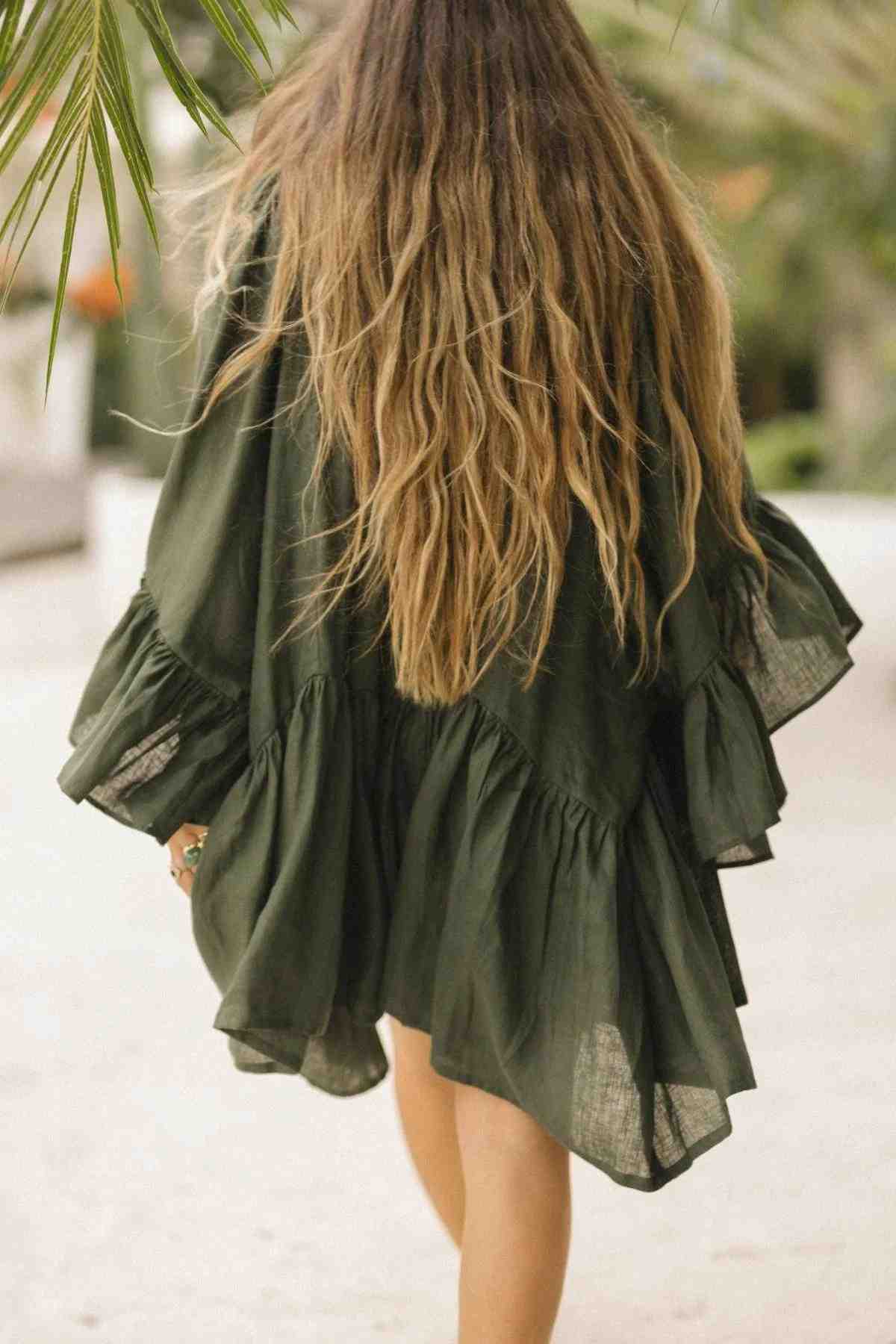 A person with long, wavy hair walks away in a dark green, loose-fitting Linen Brida Gown Short by Myrah Penaloza, embellished with ruffles. The romantic spring/summer dress drapes gracefully as they move outdoors, enveloped by the gentle whisper of green leaves in the background.