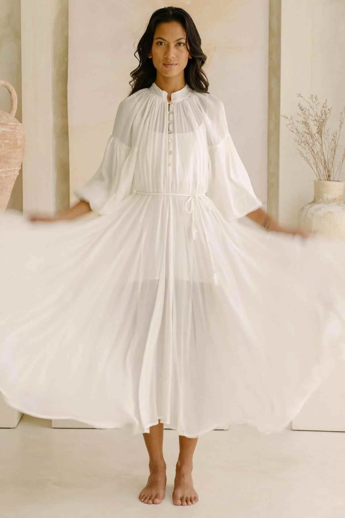 A woman stands indoors wearing a long, flowing Paloma Silk Gown from Myrah Penaloza. The bridal-inspired dress is semi-transparent with a buttoned neckline and gathered sleeves, cinched at the waist with a tie. She is barefoot, holding the sides of the dress out to create a wide, airy appearance.