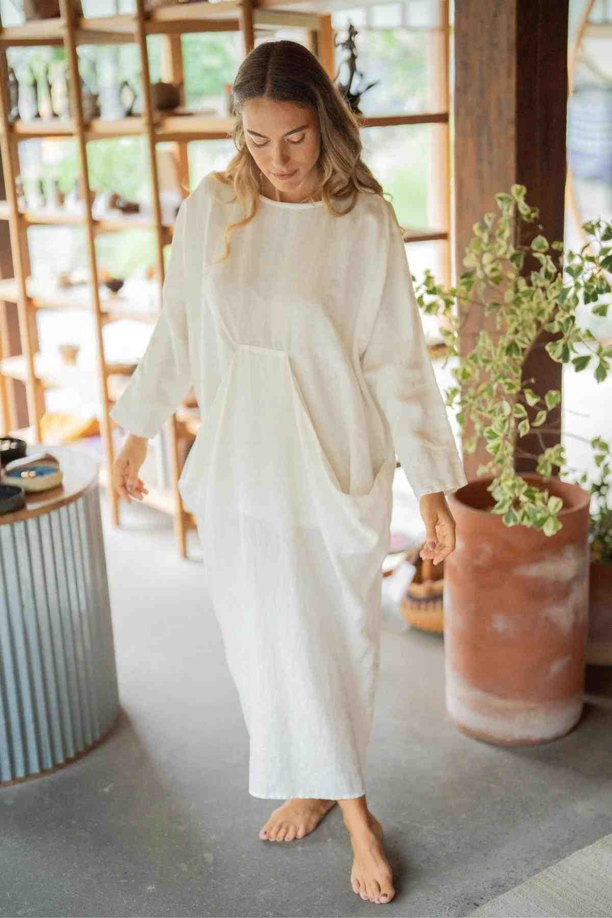 A woman in a Mountain Magic Linen Gown from Myrah Penaloza stands barefoot on a concrete floor. She is in a bright room with wooden shelves filled with various items in the background. A tall potted plant is beside her, and she looks down with a relaxed expression.