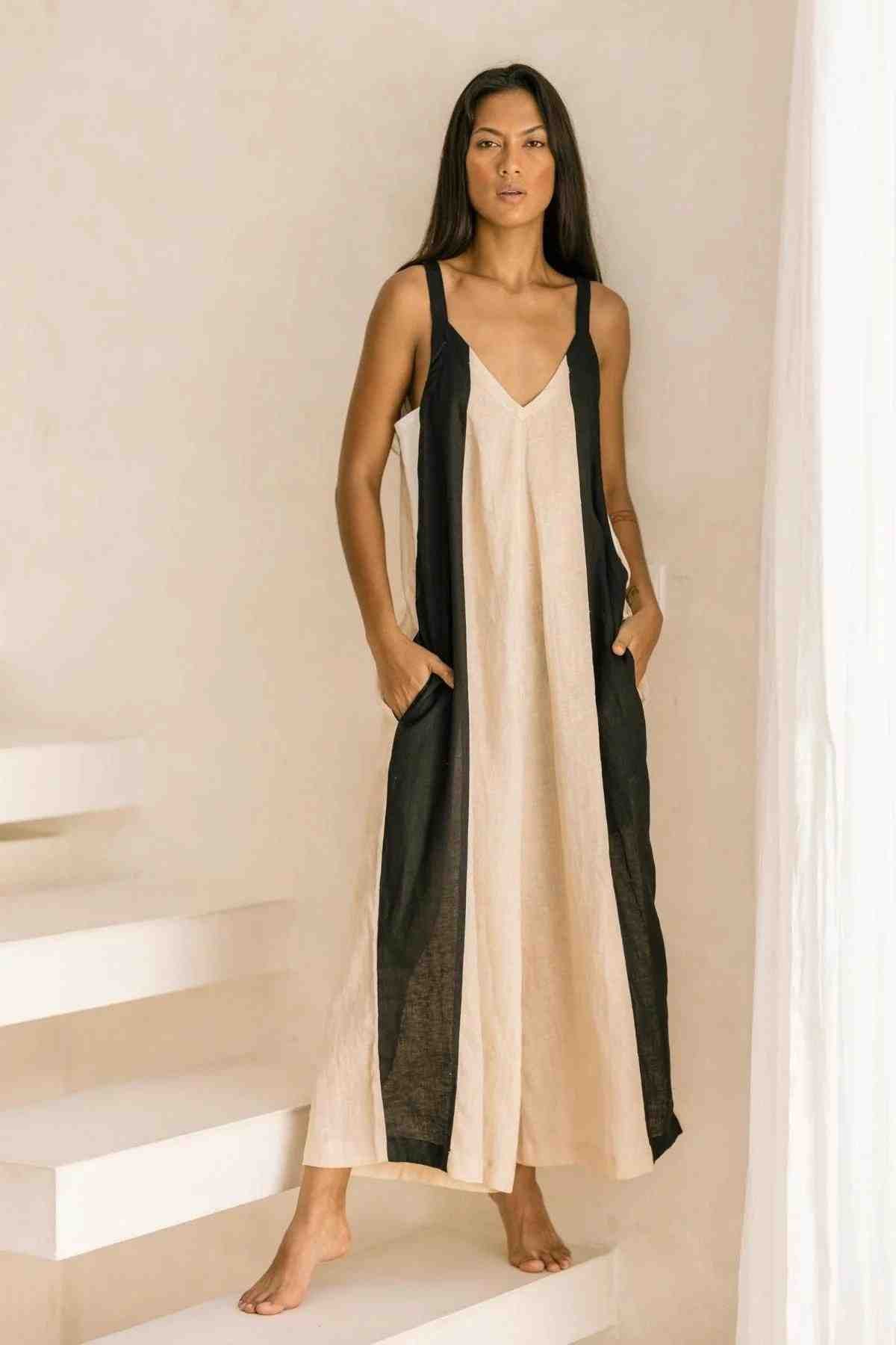 A woman stands on white steps, facing forward and wearing the Kundalini Linen Playsuit from Myrah Penaloza. The sleeveless, loose-fitting one-piece is made from luxuriously soft stonewashed linen in beige and black. Her hands rest casually in the playsuit’s pockets, and she gazes directly at the camera against a plain, light-colored background.