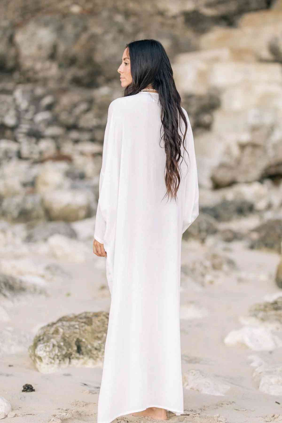 A woman with long dark hair stands on a rocky beach, facing away and wearing a flowing white Bamboo Rayon Virgo Rising Gown by Myrah Penaloza. The background is comprised of weathered stones and sand. She appears contemplative, adding a serene atmosphere to the scene.
