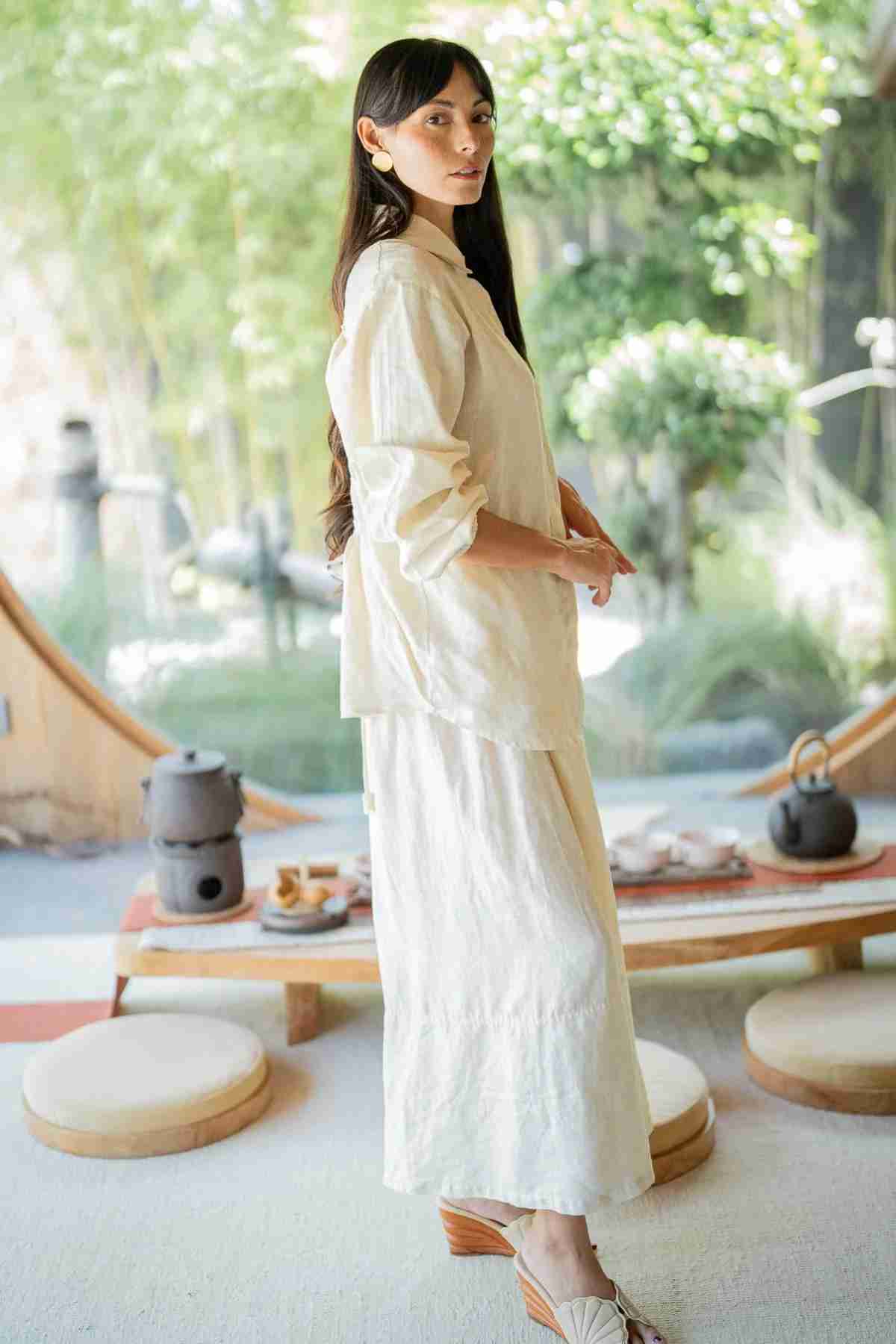 A woman with long dark hair is indoors, donning an oversized outfit that includes the Pantalon Linen Pants (Wide Legged) by Myrah Penaloza. Behind her, a tea set sits on a wooden table, and greenery is visible through a large window, contributing to the serene atmosphere.