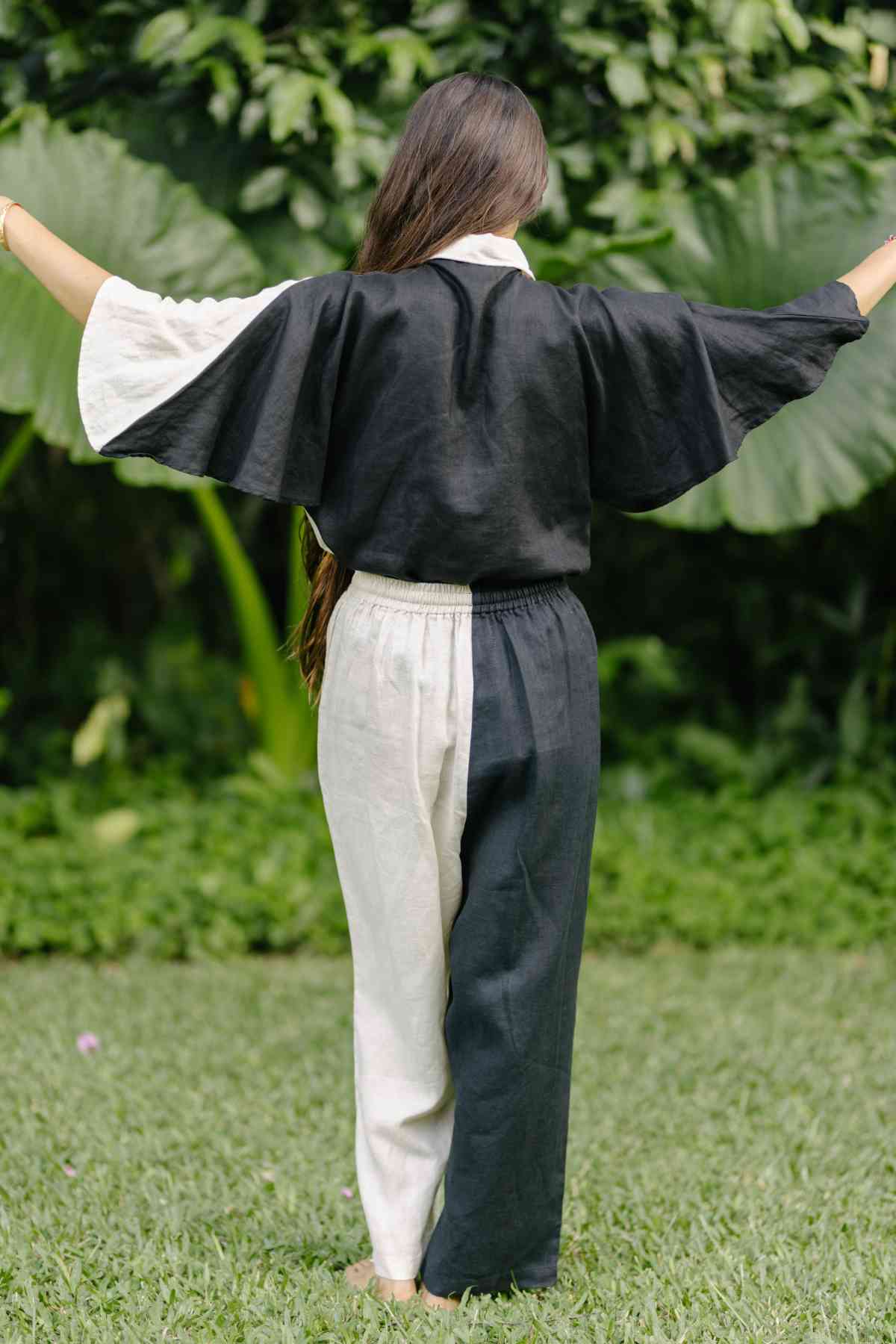 A person stands with arms outstretched, facing away from the camera, wearing a Linen Kiki Yin Yang Set (Made To Order) by Myrah Penaloza. The outfit is made of 100% stone-washed linen and includes a top and matching pants. The person is barefoot against a backdrop of lush green plants and grass.