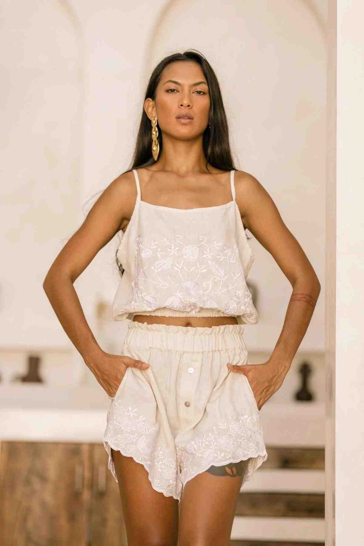 A woman stands confidently wearing the Myrah Penaloza Wonder Sleeper Set in stonewashed embroidered linen, which includes a hand-embroidered floral crop top and matching shorts. The background showcases a light, neutral-toned interior with soft arches. Her long, straight hair complements the look, accented by a single large earring.