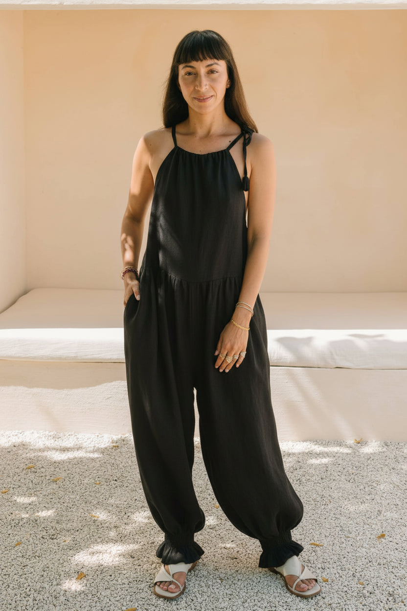 Front view of a woman standing in the Monaya Playsuit by Myrah Penaloza in Dark Moon black stone washed linen. Oversized, full length silhouette with tie shoulder straps, hand in pocket, handcrafted in Bali.