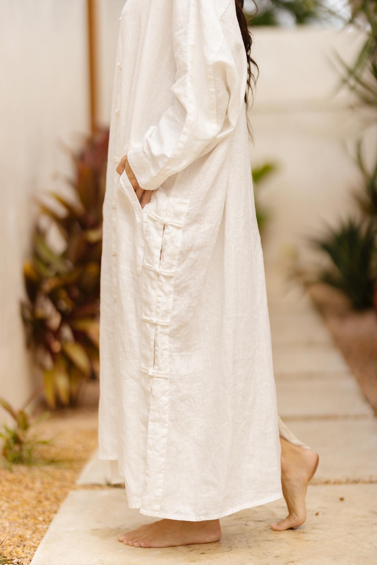 A person in the Ancient Chajin Jacket (100% Linen) from the Heaven & Earth Collection by Myrah Penaloza stands barefoot on a stone pathway. Their hands are in the pockets of the jacket. The background includes plants with green and reddish-brown leaves, creating an outdoor and sunlit setting.