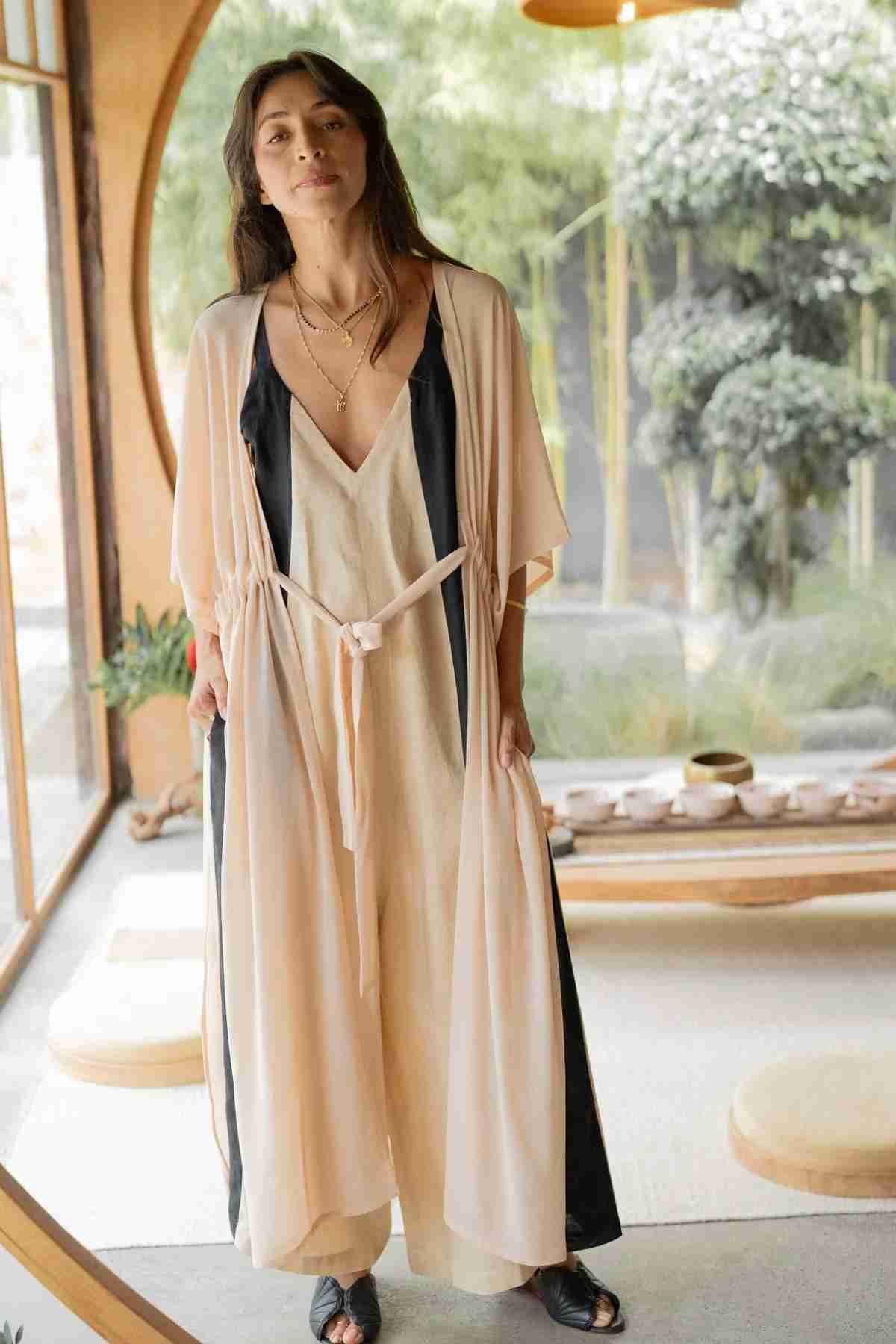 A woman stands indoors wearing a Myrah Penaloza Madelin Sheer Chiffon Long Cover with an elegant, light-colored design and a dark inner lining that exudes sheer brilliance. She holds the sides of her cover with her hands. Near a window showcasing outdoor greenery, a wooden table adorned with teacups and saucers can be seen in the background.