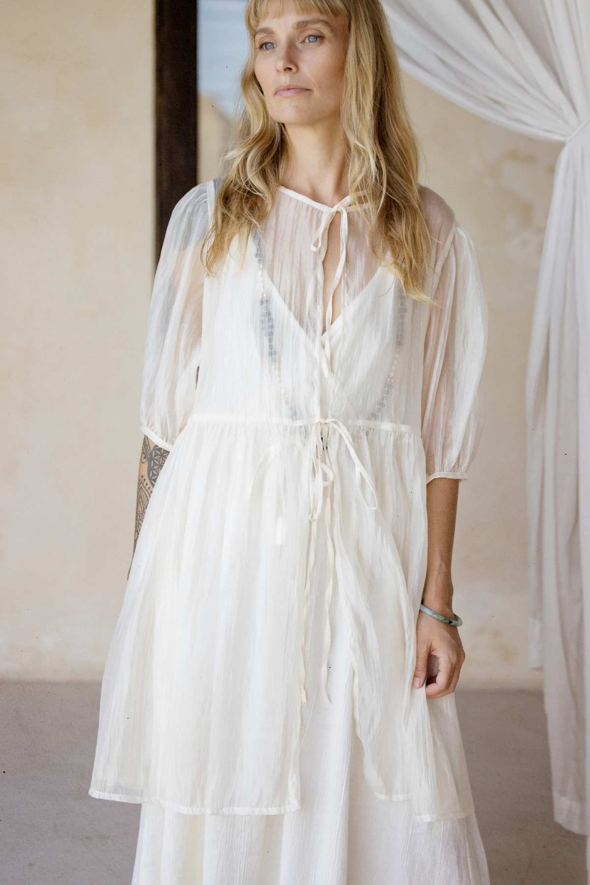 A woman dressed in a sheer, flowing Dora Slip Dress from Myrah Penaloza stands indoors, radiating effortless elegance as she gazes to the side. This timeless piece boasts a tie at the neckline and layered fabric. The softly lit environment, complemented by neutral-toned walls and curtains, enhances its charm.