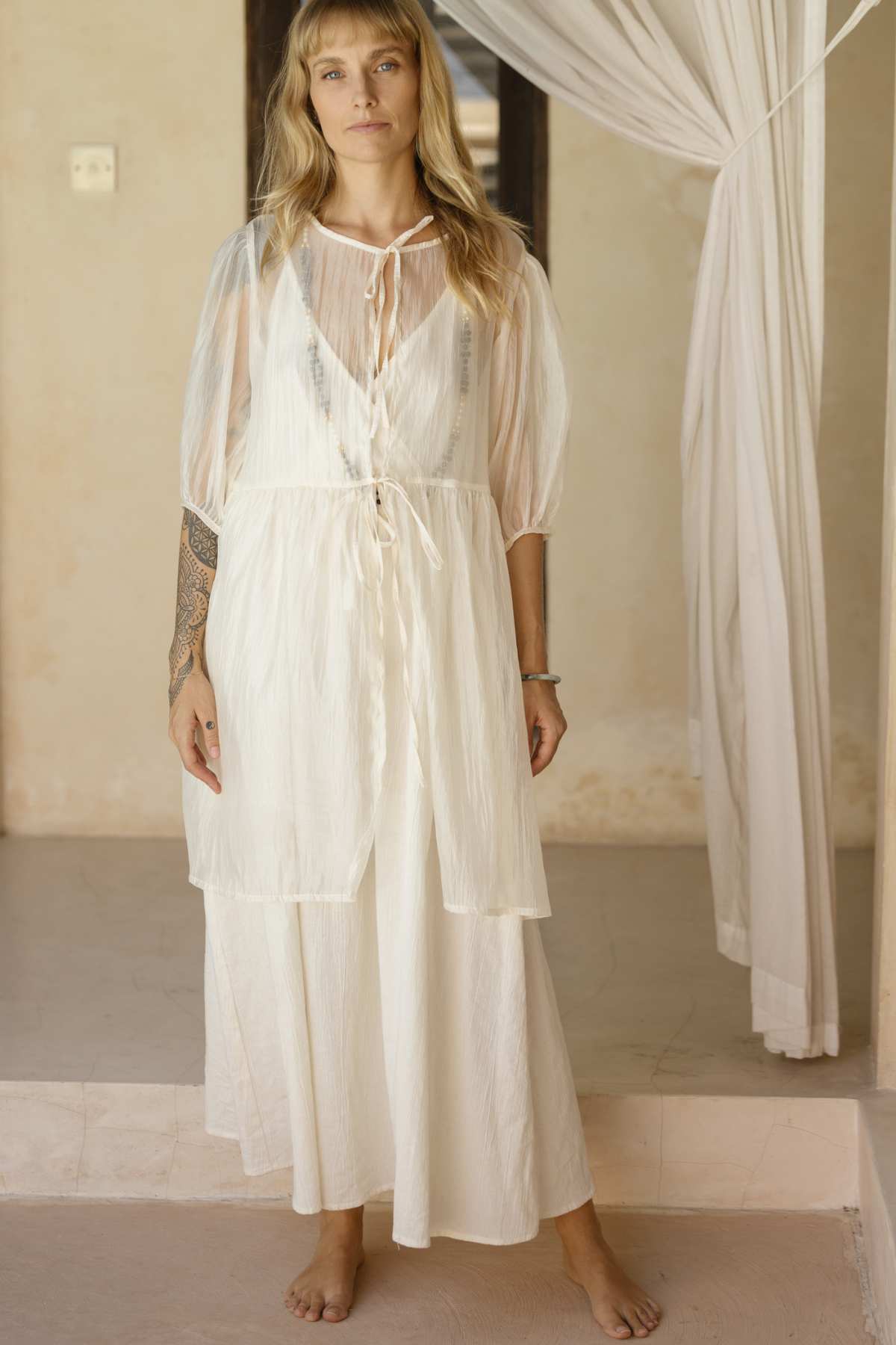 A woman stands barefoot indoors, radiating effortless elegance in a sheer, flowing white Dora Slip Dress by Myrah Penaloza, layered over a longer gown. Her hair cascades down, revealing a tattoo on her right arm. The setting is timeless, with neutral tones and softly draped curtains in the background.