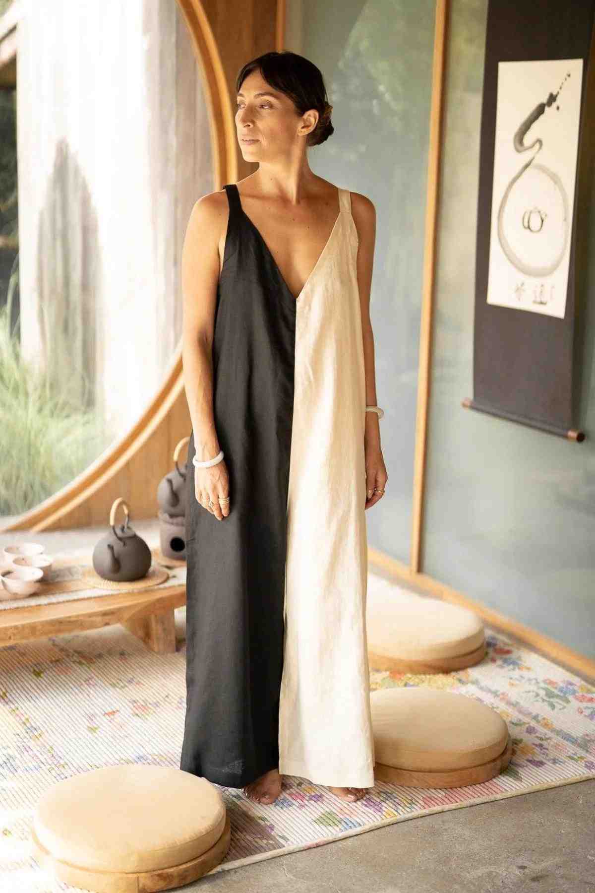 A woman stands in a minimalist room, dressed in the Kundalini Linen Playsuit by Myrah Penaloza—a flowing, two-toned ensemble with black on one side and beige on the other. The luxuriously soft stonewashed linen drapes elegantly as she looks to her left, barefoot. The room features a circular window, meditation cushions, a tea set, and an art scroll on the wall.