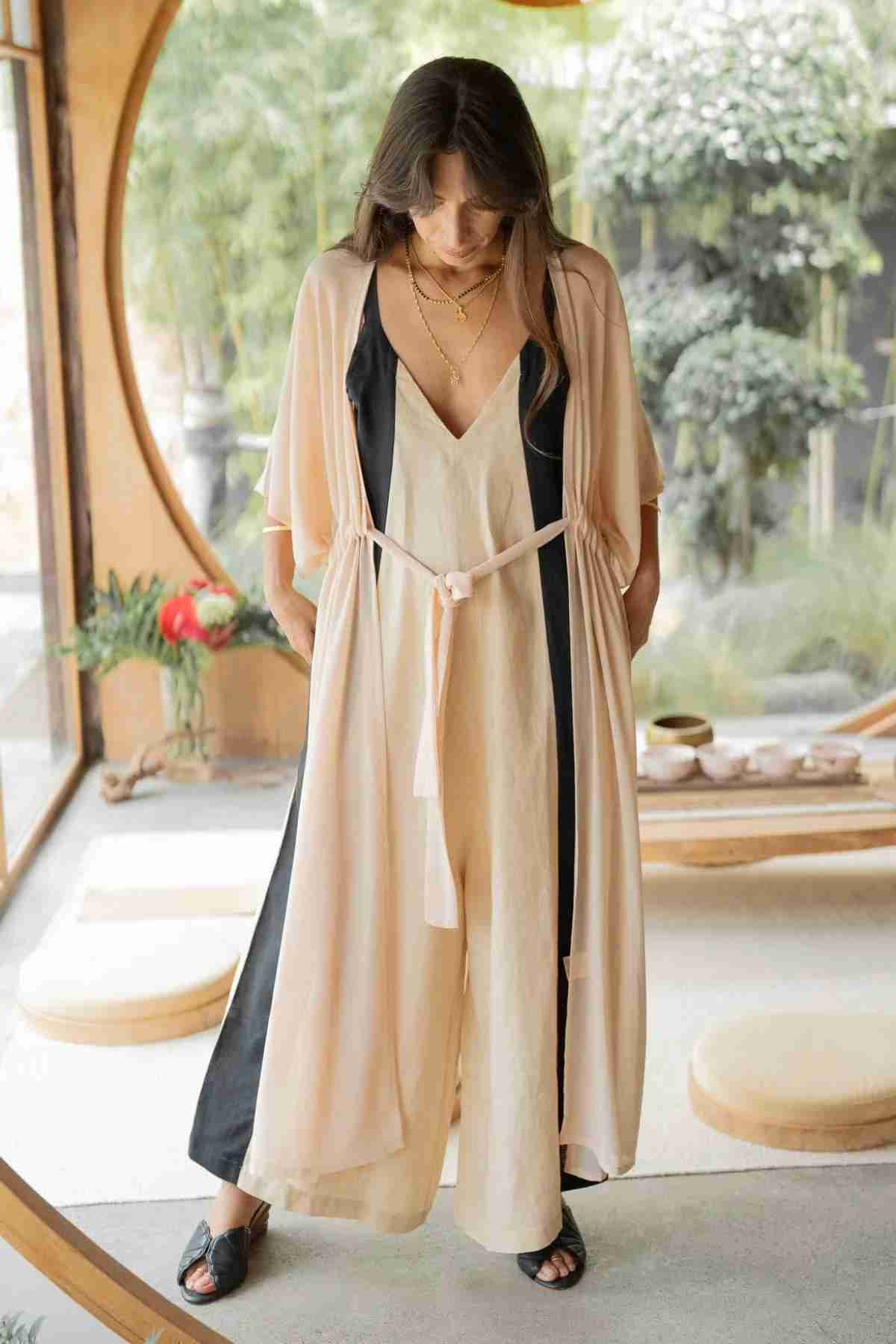 A woman stands in a serene, minimalistic room with large circular windows overlooking greenery. She is wearing a light, flowing sleeveless dress paired with a Madelin Sheer Chiffon Long Cover by Myrah Penaloza, tied elegantly at the waist and complemented by black sandals. Her head is slightly bowed, and her hands are placed in her pockets.