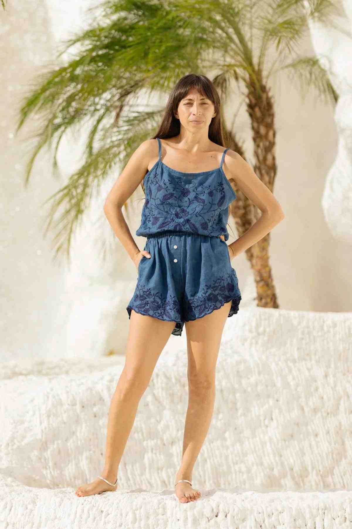 A woman in an intricately designed romper, reminiscent of the Wonder Sleeper Set by Myrah Penaloza, stands confidently on a textured white surface, bordered by palm trees and a striking white cliff. Her ensemble is complemented by white sandals and her long hair cascading over her shoulders.