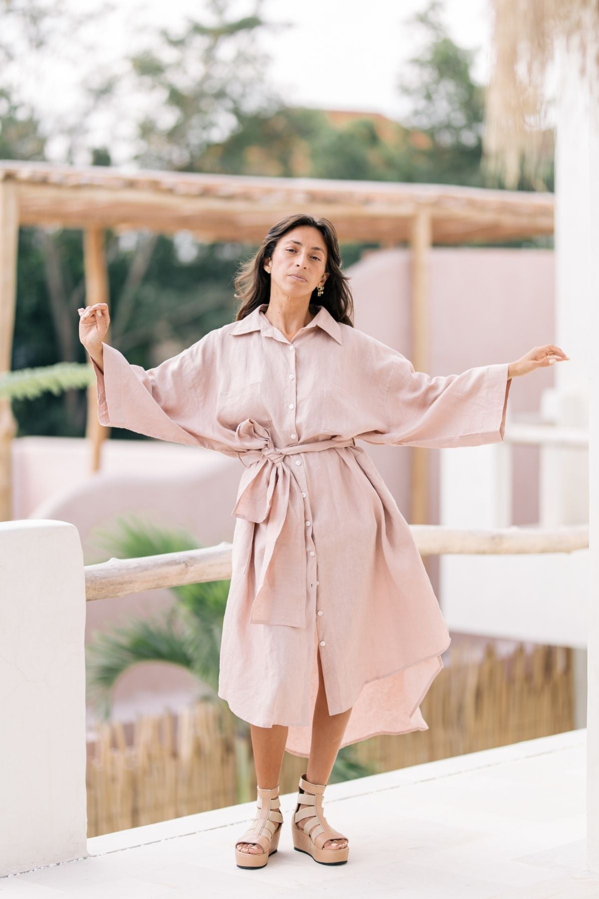A woman stands on a balcony with light pink walls and wooden elements in the background. She is wearing the Nidra Full Length Linen Duster by Myrah Penaloza, a breathable, light pink dress with a tie belt and beige strappy wedge sandals. The oversized fit offers multiple styling options, while the background vegetation indicates a serene, outdoor setting.